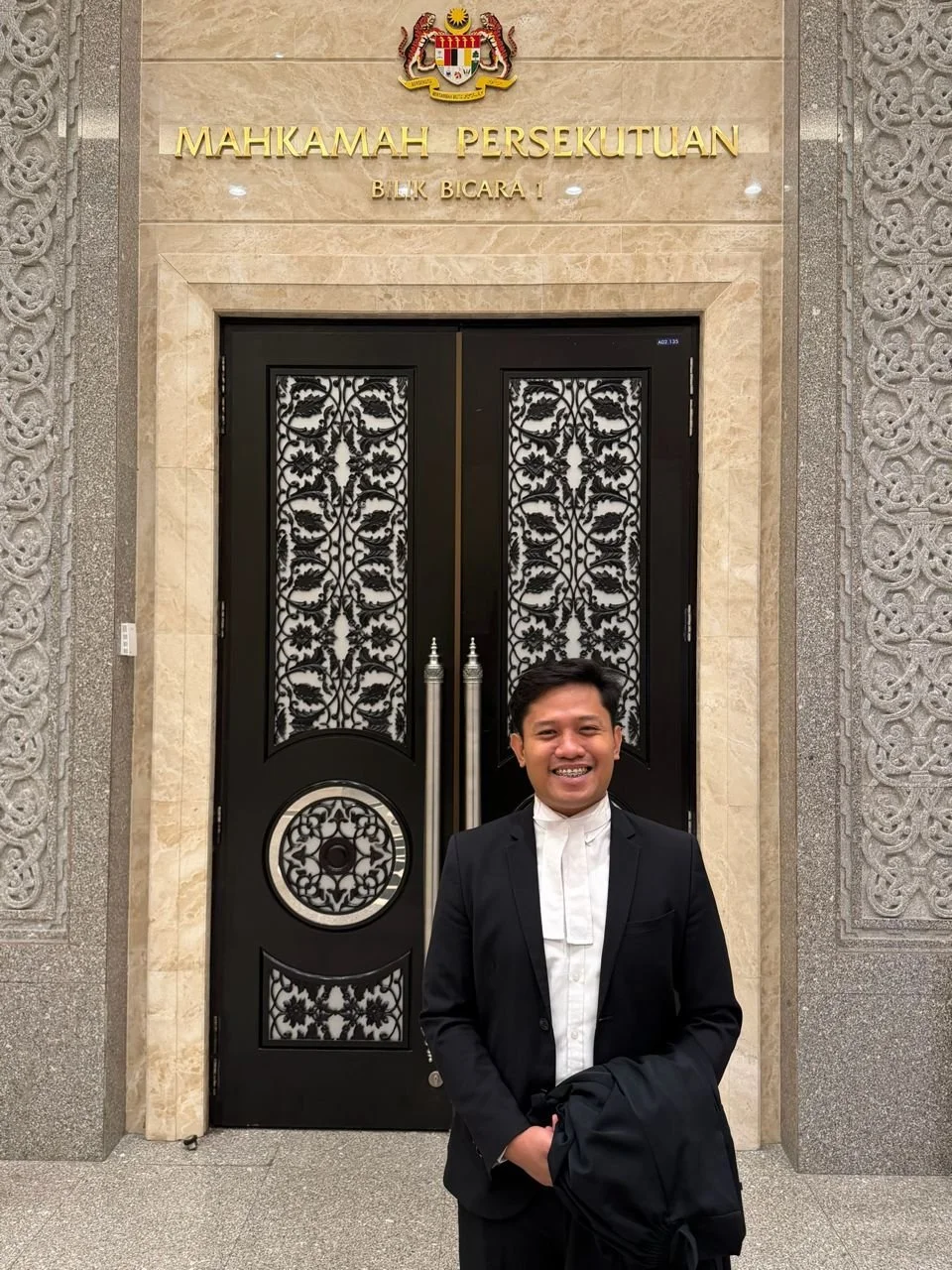 A smiling man in a black suit and white shirt standing in front of an ornate black door with intricate metalwork. The background features a beige wall with gold lettering and the emblem above the door, indicating the Court of Appeal in Malaysia.