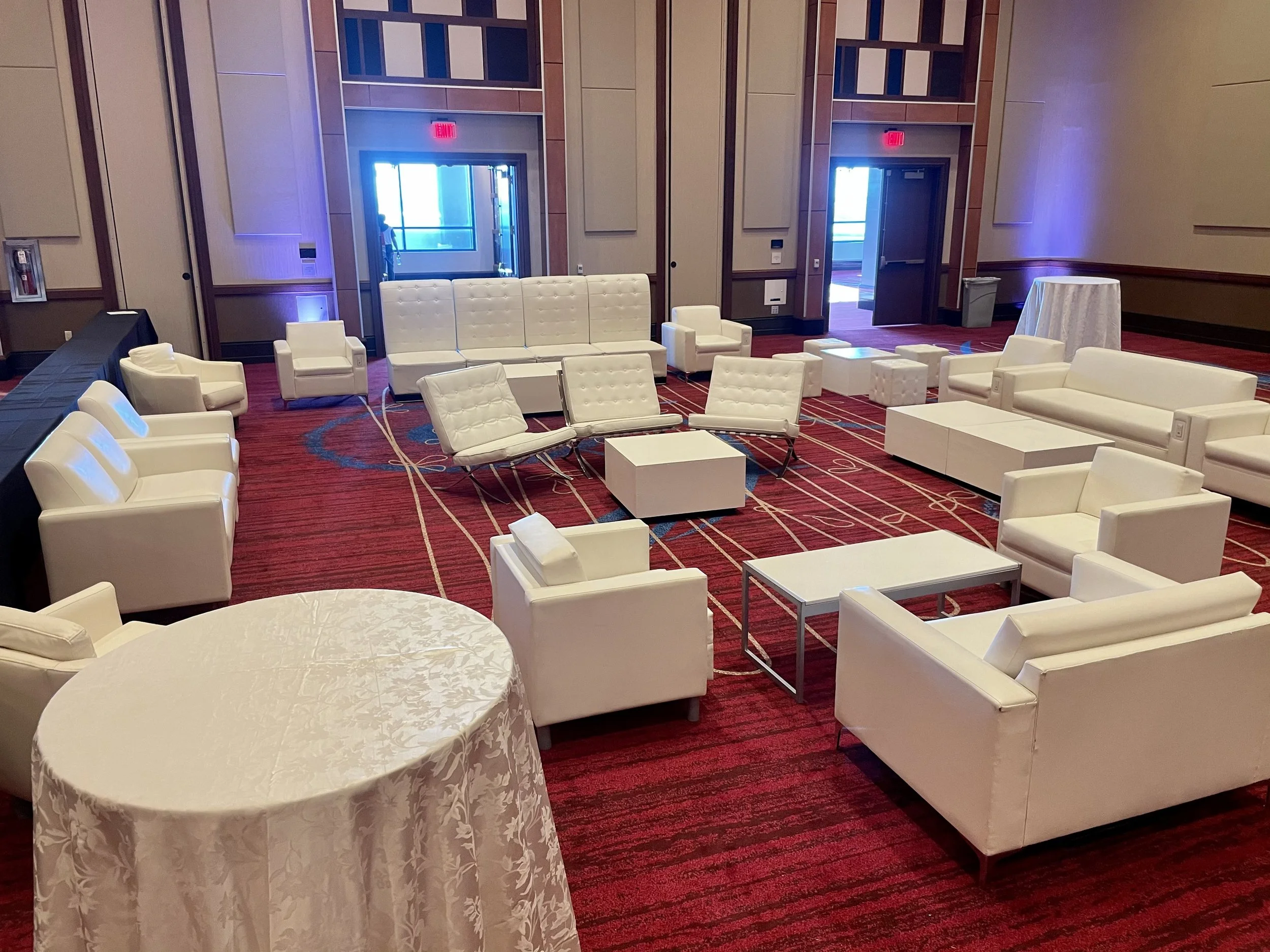 A spacious lounge area with white modern couches and armchairs arranged around white tables. A round table with a white tablecloth is in the foreground. The room has red and blue patterned carpet and wooden paneling on the walls. Double doors with exit signs are visible in the background, with a person seen through one of the doors.