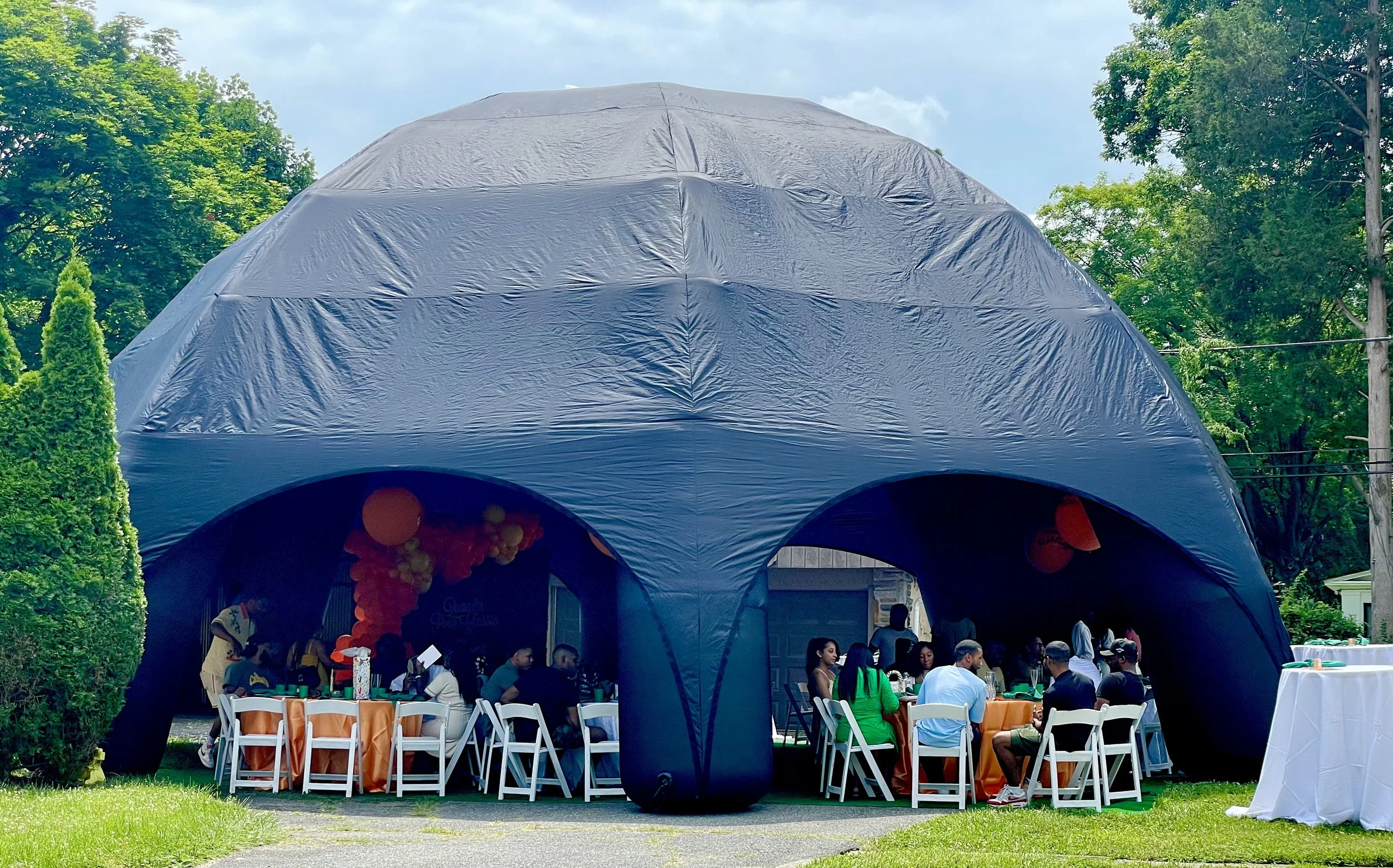 A large blue event tent set up in a residential area with people gathered underneath, surrounded by greenery and trees, with some tables and chairs inside and outside the tent.