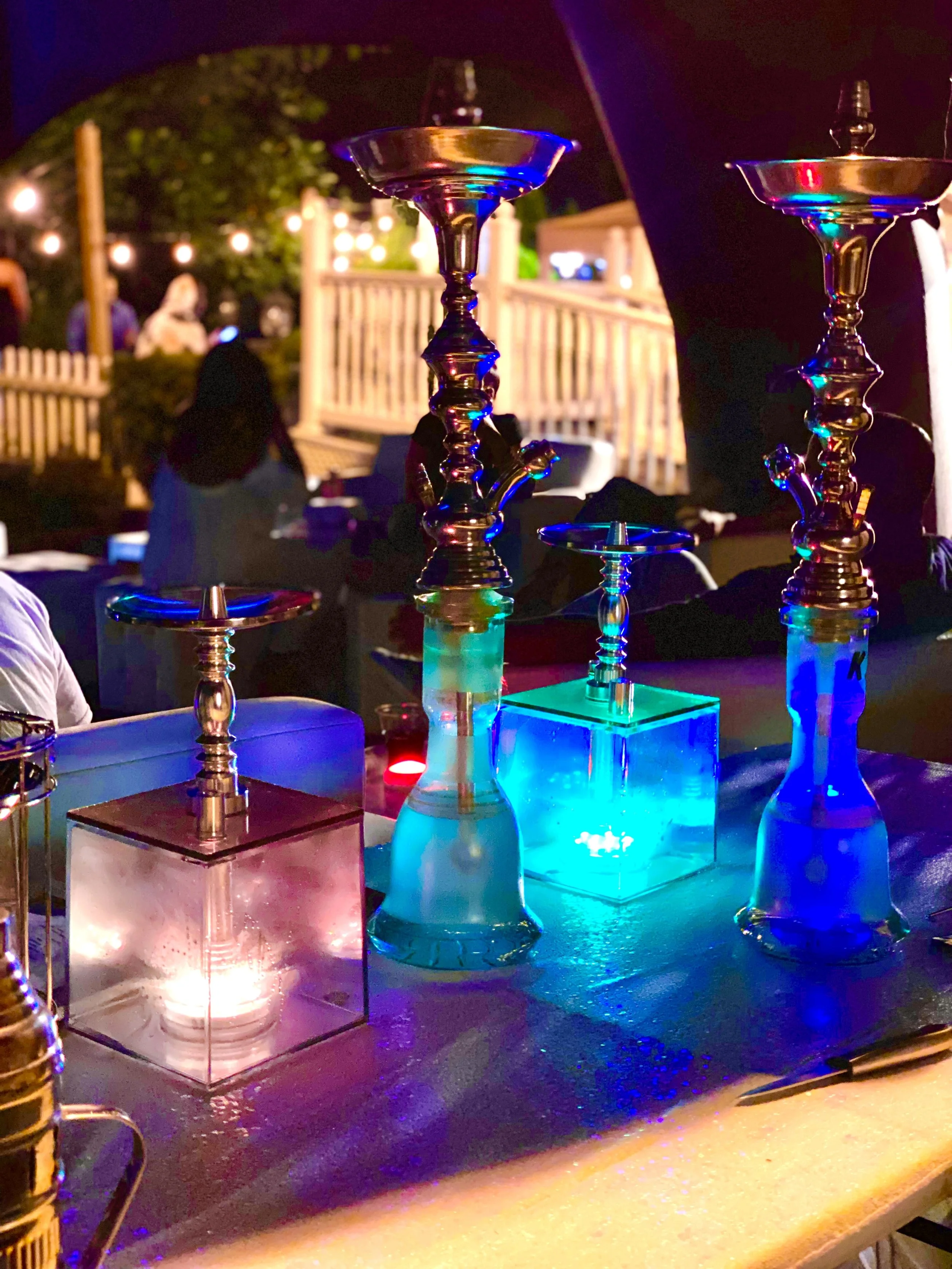 Colorful hookah pipes on a table illuminated with vibrant blue, pink, and purple lights, with blurred people and string lights in the background at night.