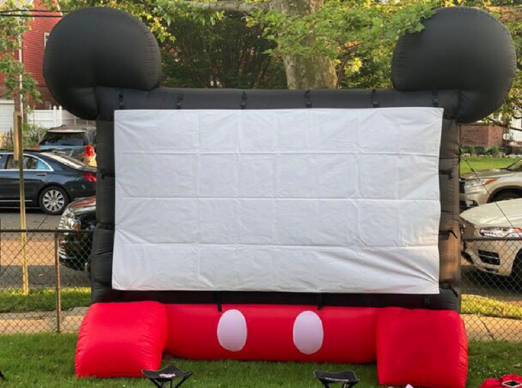 Inflatable movie screen setup with a Mickey Mouse theme, featuring large black ears and a red base with white buttons, set outdoors on a grassy area with cars and trees in the background.