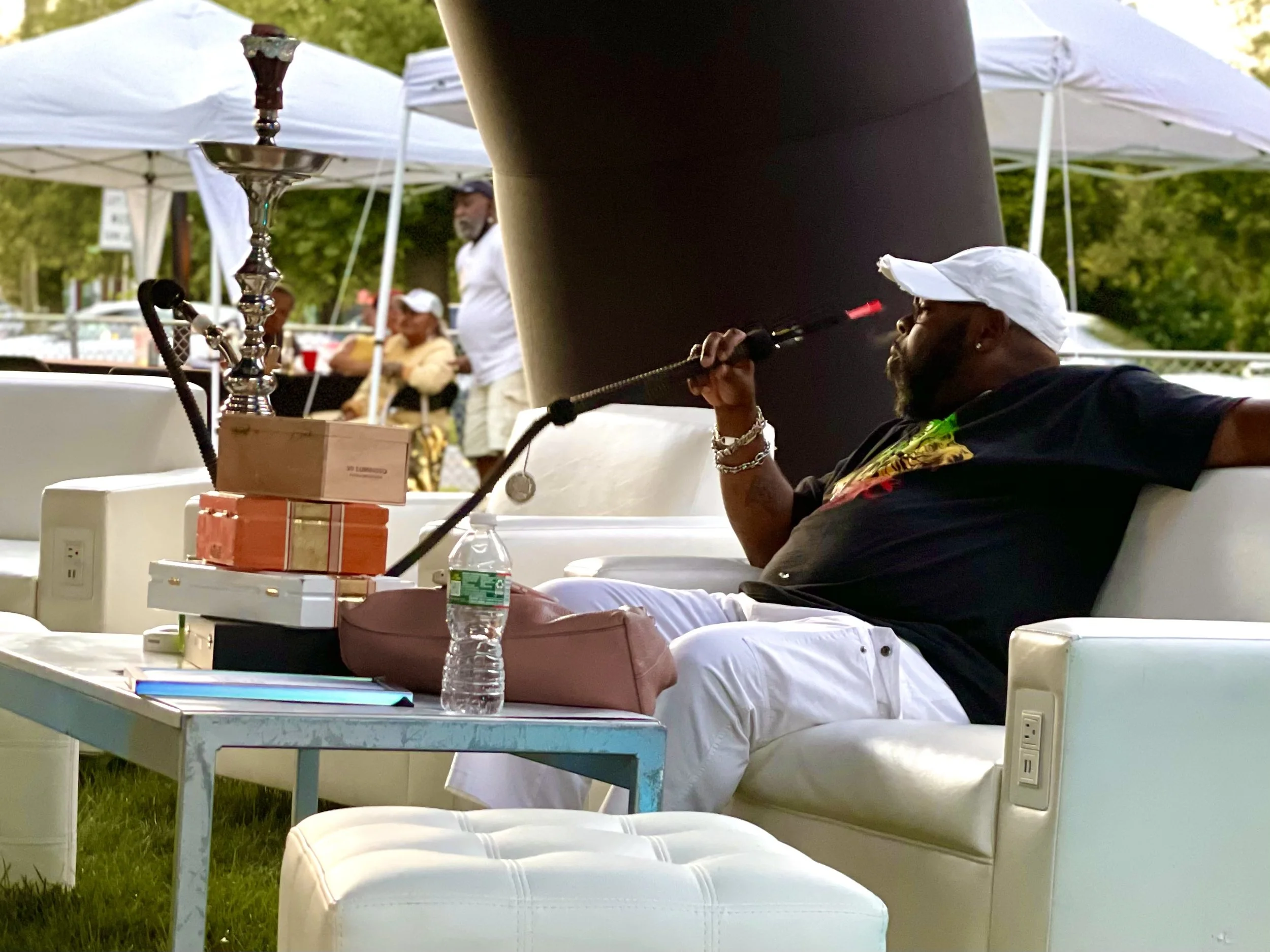 A person with a white cap and black shirt using a hookah at an outdoor event with white tents and several people seated in the background.