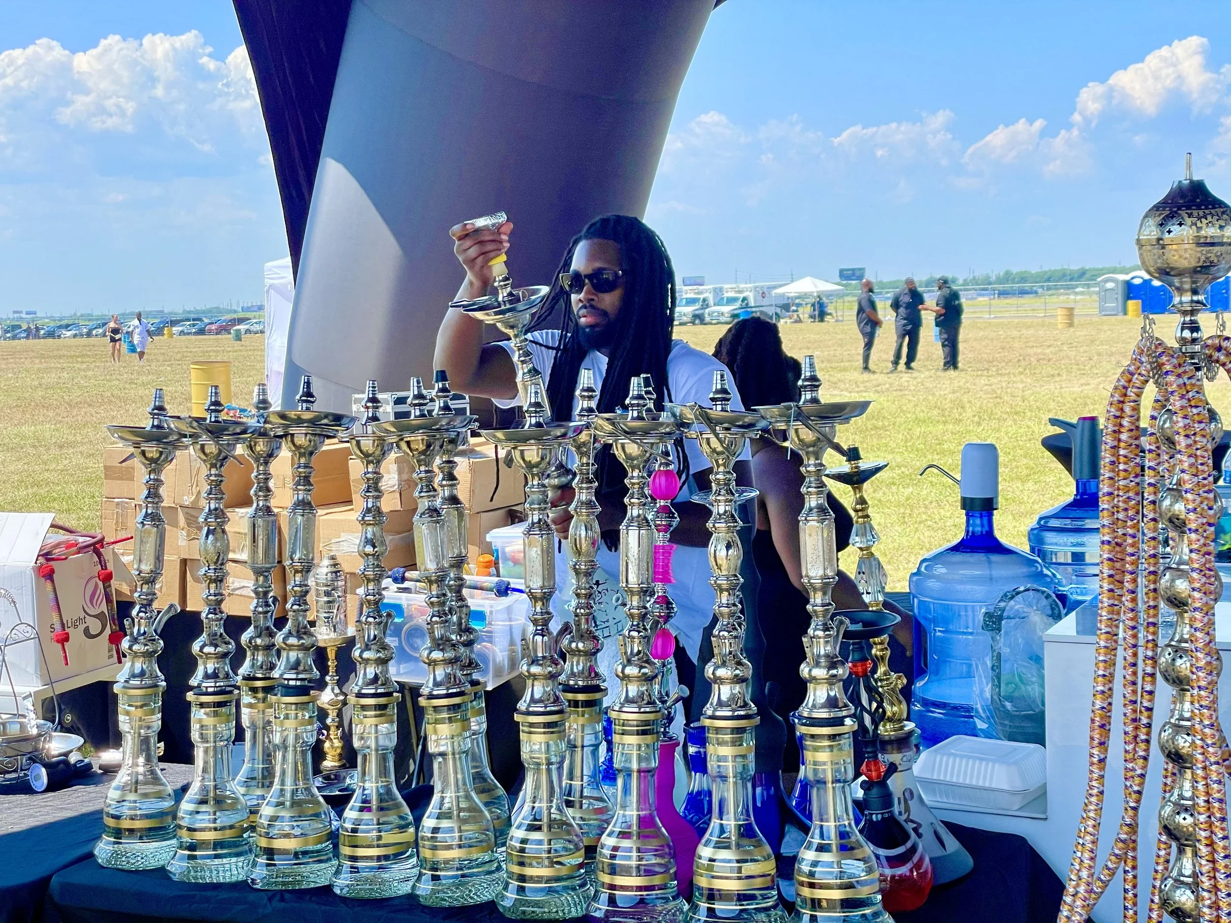 A man with long dreadlocks wearing sunglasses standing behind a table of hookah pipes at an outdoor event. The table is filled with various glass hookah bases, and there are other items like bottles and accessories. The background shows a wide grassy field with a few people walking and some tents under a blue sky with scattered clouds.