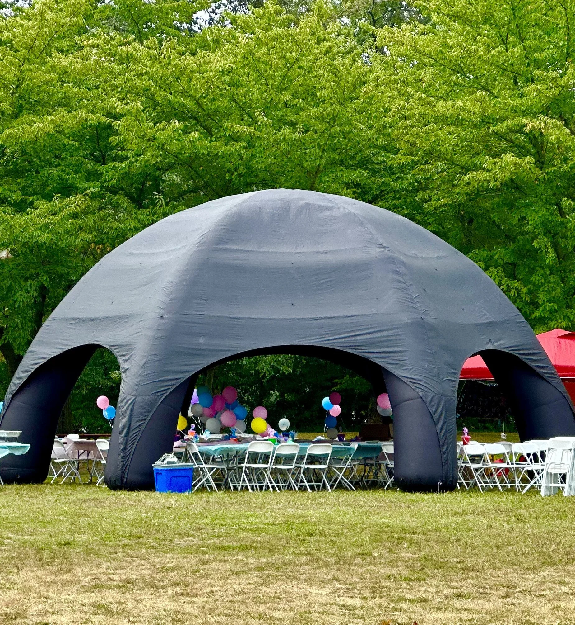 Outdoor celebration with tables, chairs, balloons, and a large inflatable tent. Green trees are in the background.