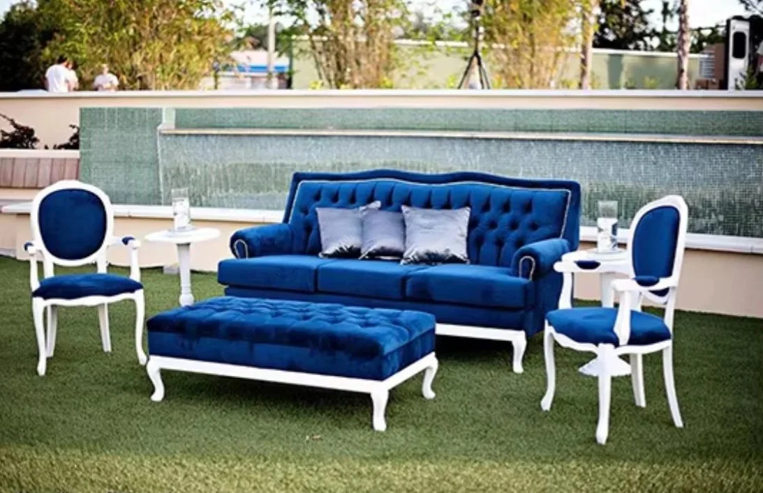 Blue outdoor sofa with matching ottoman and two matching armchairs with white frames on a grassy area, with small white tables, and a water feature in the background.
