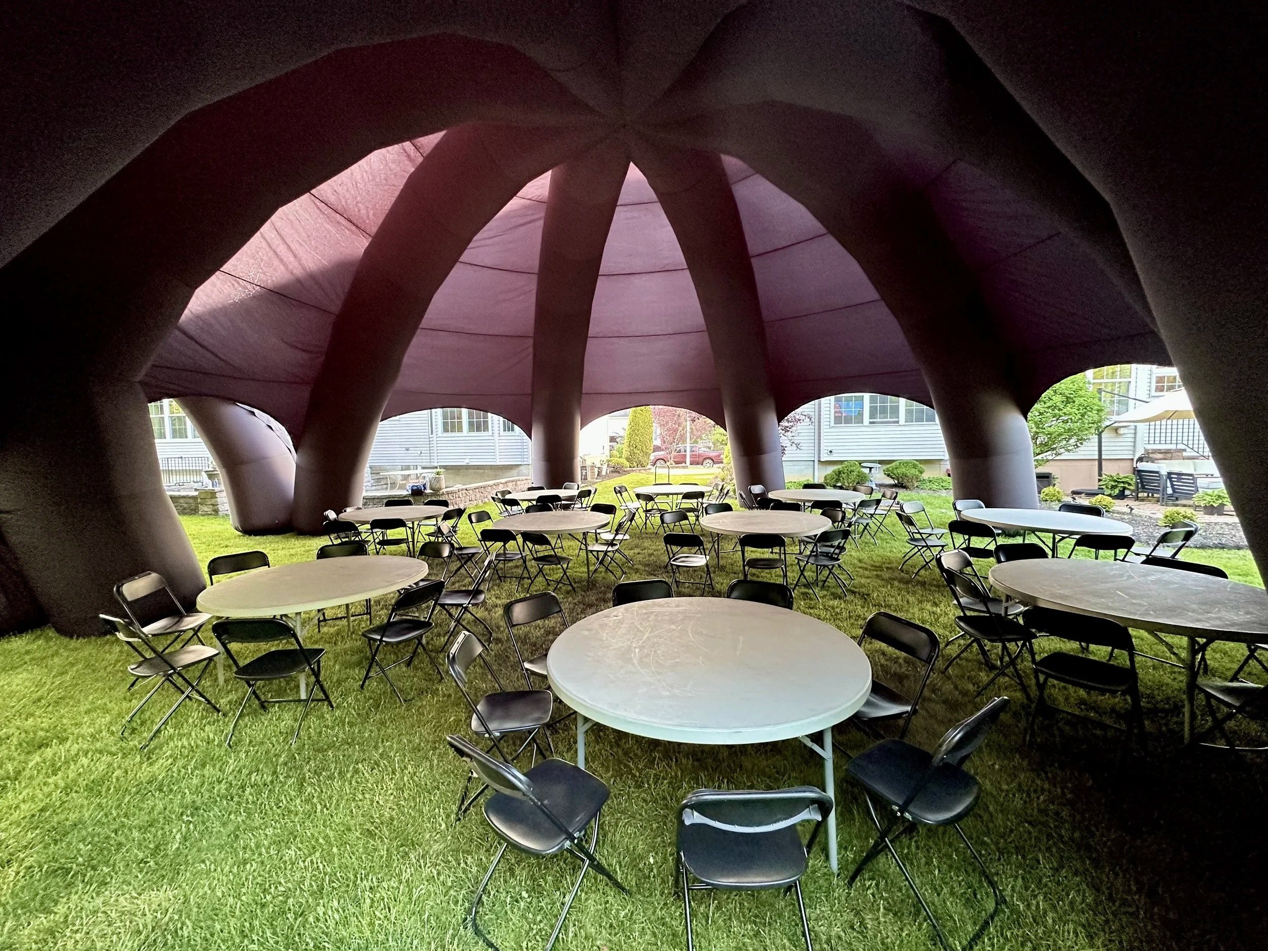 Under a large, pink, tent-like structure with supporting thick, tentacles, there are multiple round tables with black chairs on green grass.