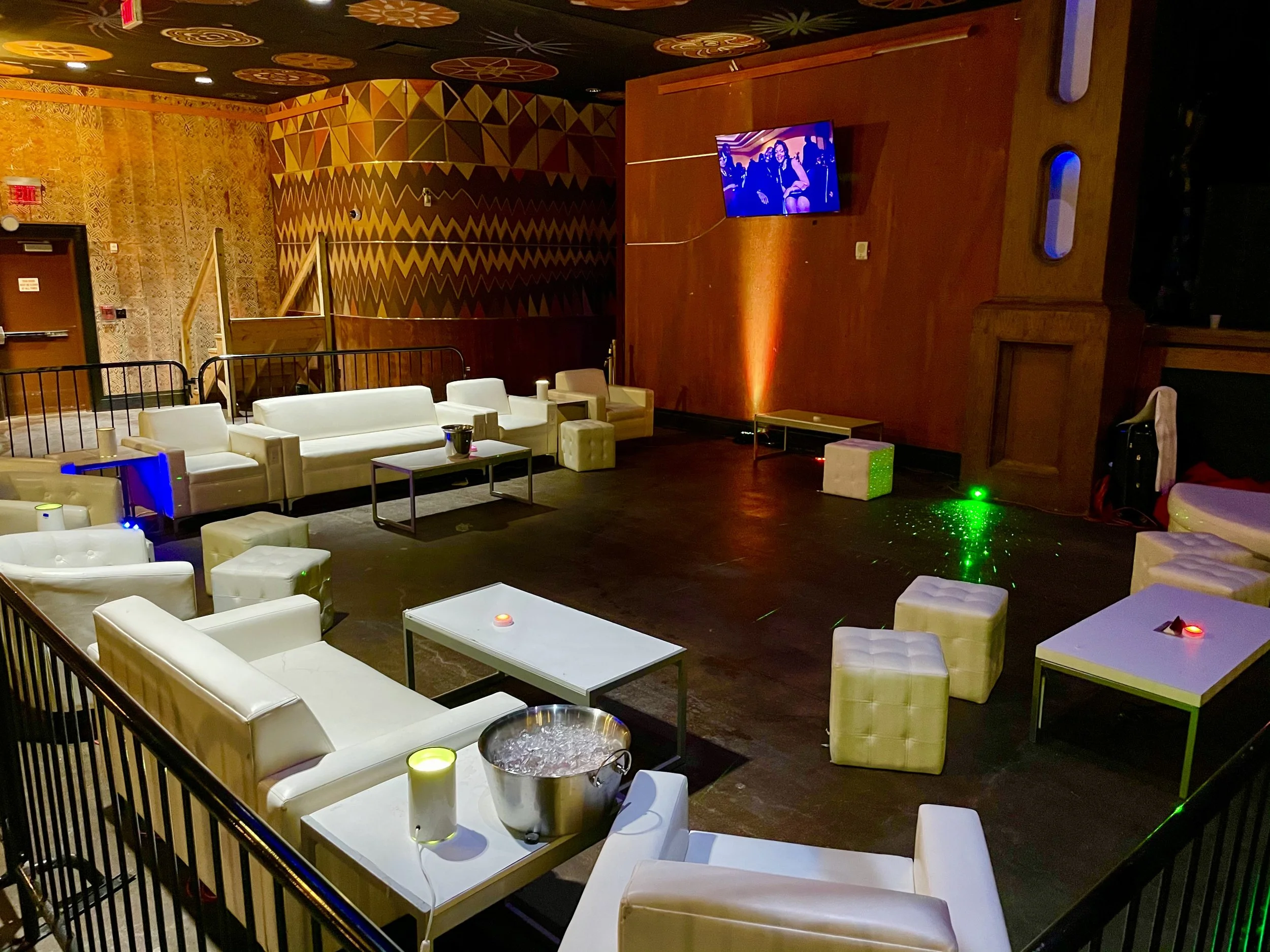An indoor lounge area with white sofas, small tables, and colorful decorative wall and ceiling patterns, with a mounted television and some DJ or lighting equipment on the stage.