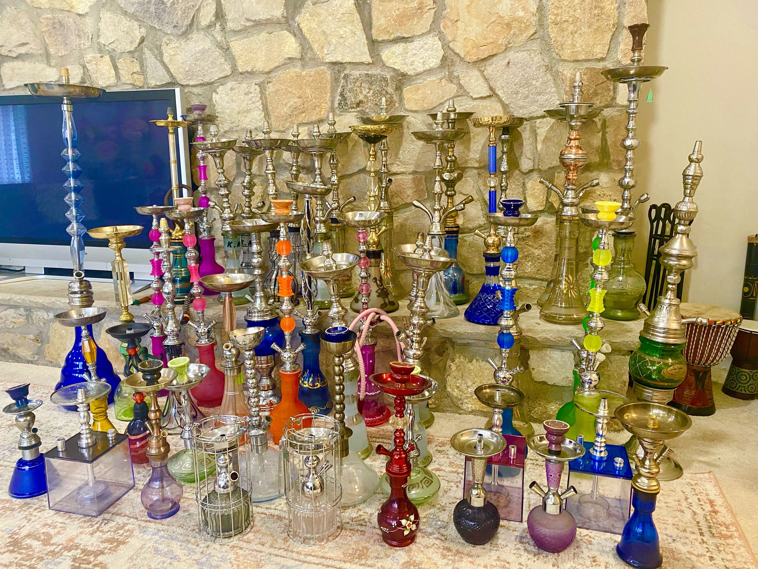 A collection of colorful glass and metal hookahs on a table against a stone wall.
