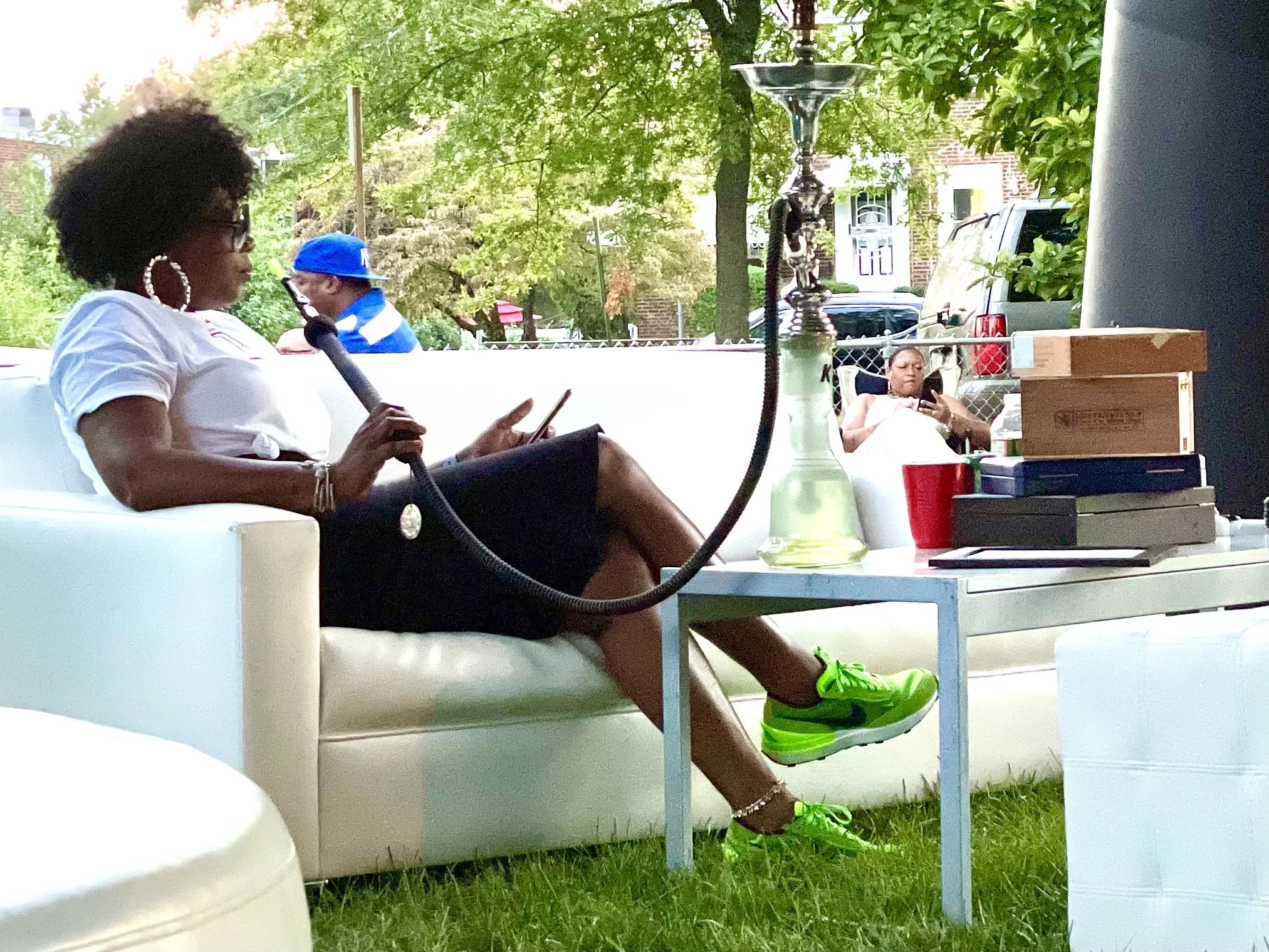 A woman with black curly hair, glasses, and large hoop earrings is sitting on a white outdoor couch, looking at her phone. She is wearing a white shirt, black shorts, and bright green shoes. There are outdoor plants and trees in the background, with a woman in a white dress sitting at a table and looking at her phone.