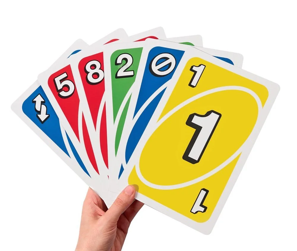 A hand holding a set of large, colorful Uno cards fanned out, showing the numbers 1, 2, 5, 8, and special cards with symbols and numbers in blue, red, green, and yellow.