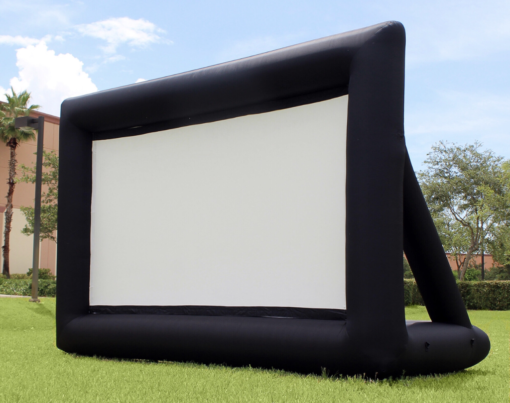 An inflatable movie screen set up outdoors on a grassy area with trees and buildings in the background under a blue sky.
