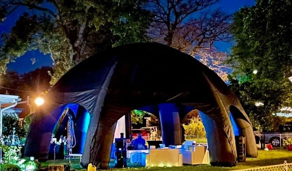 An outdoor nighttime scene with a large black inflatable tent or dome, illuminated in blue and yellow, set up in a backyard with trees, outdoor furniture, and string lights.