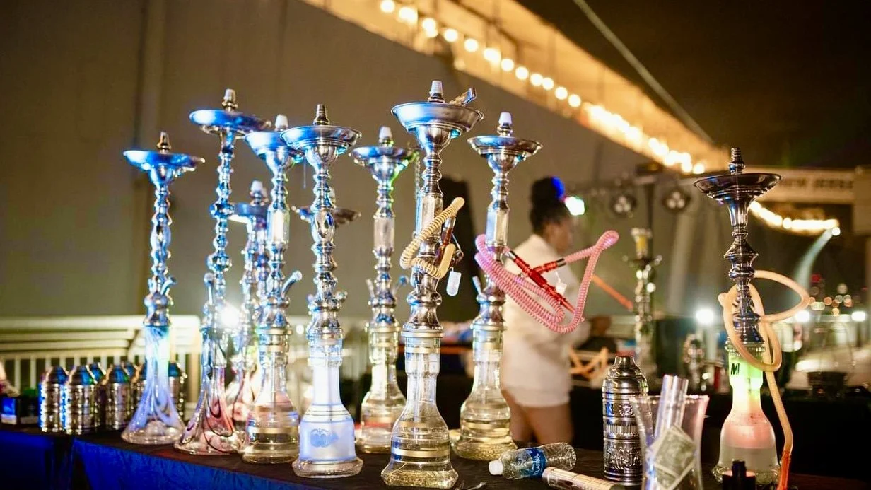 Multiple hookah pipes with colorful hoses on a table at a hookah lounge, with a woman in the background.