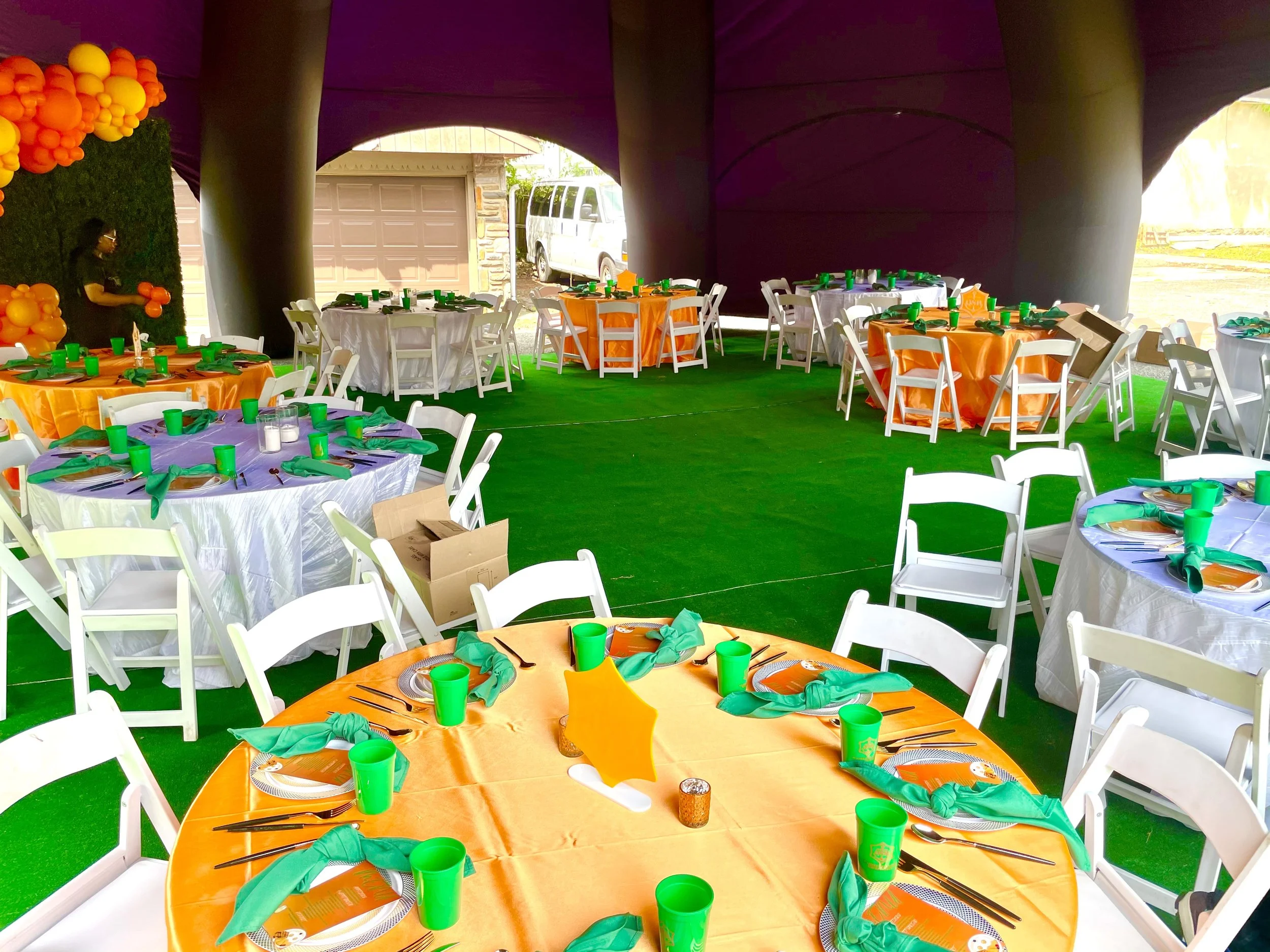 Decorated party space with round tables covered in orange and white tablecloths, set with green cups, green napkins, and utensils. Round tables are arranged on green artificial turf, with white chairs around them. The background shows an underpass or covered area with purple fabric overhead and a view of a garage door and parked van outside.
