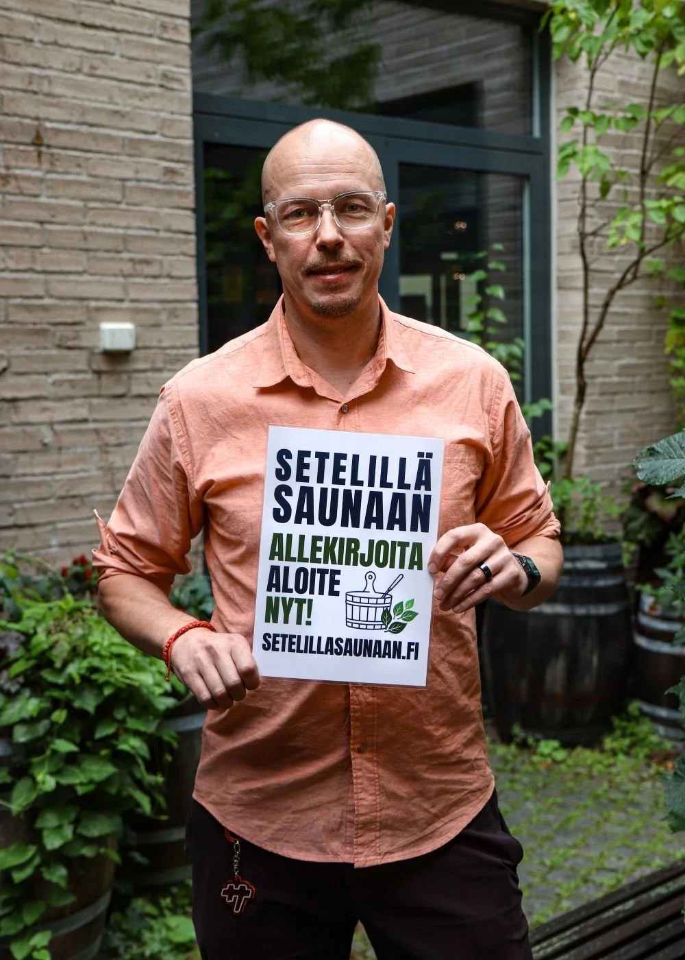 Mies seisoo puistossa, jossa on kasveja ja rakennuksen ikkuna taustalla, ja hän pitää julistetta.