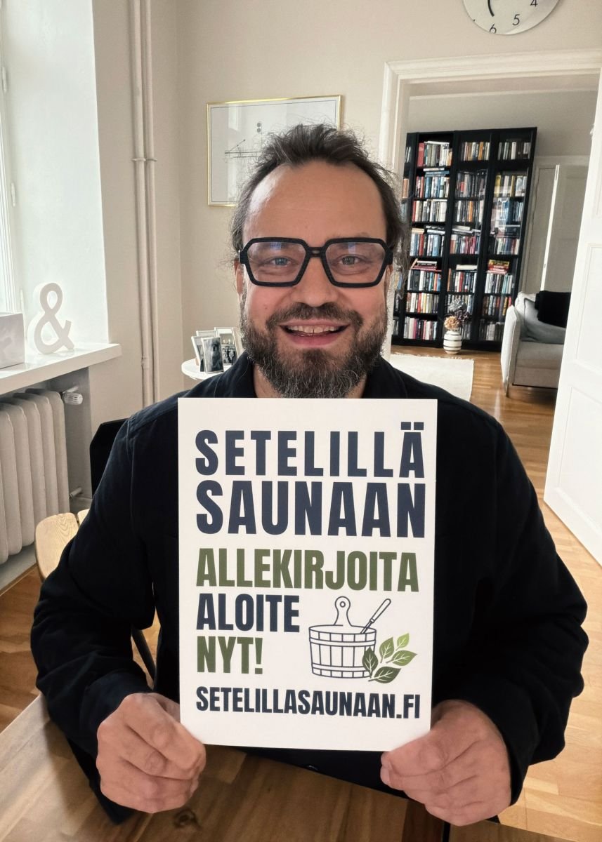 Hymyilevä mies, jolla on harmaa parta ja lasit, istuu ruokapöydällä ja pitää ylläkyltteä, jossa mainostetaan saunaa ja kannustetaan allekirjoittamaan. Taustalla on kirjahylly, pieni pöytä ja valkoinen seinä.