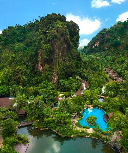 Landschaft mit steilem, bewaldetem Hügel und einem Resort mit Swimmingpools, umgeben von üppigem Grün und kleinen Gebäuden.