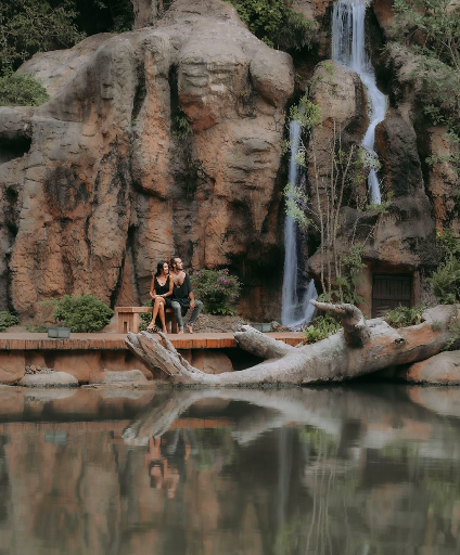 Zwei Frauen sitzen auf einer Plattform vor einem Wasserfall mit felsiger Felswand im Hintergrund, in der Nähe eines ruhigen Sees.