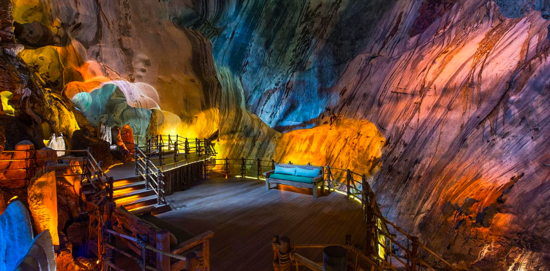 Innenansicht einer farbenfrohen Höhle mit Holzbrücke, Sitzgelegenheit und beleuchteten Wänden in verschiedenen Farben.