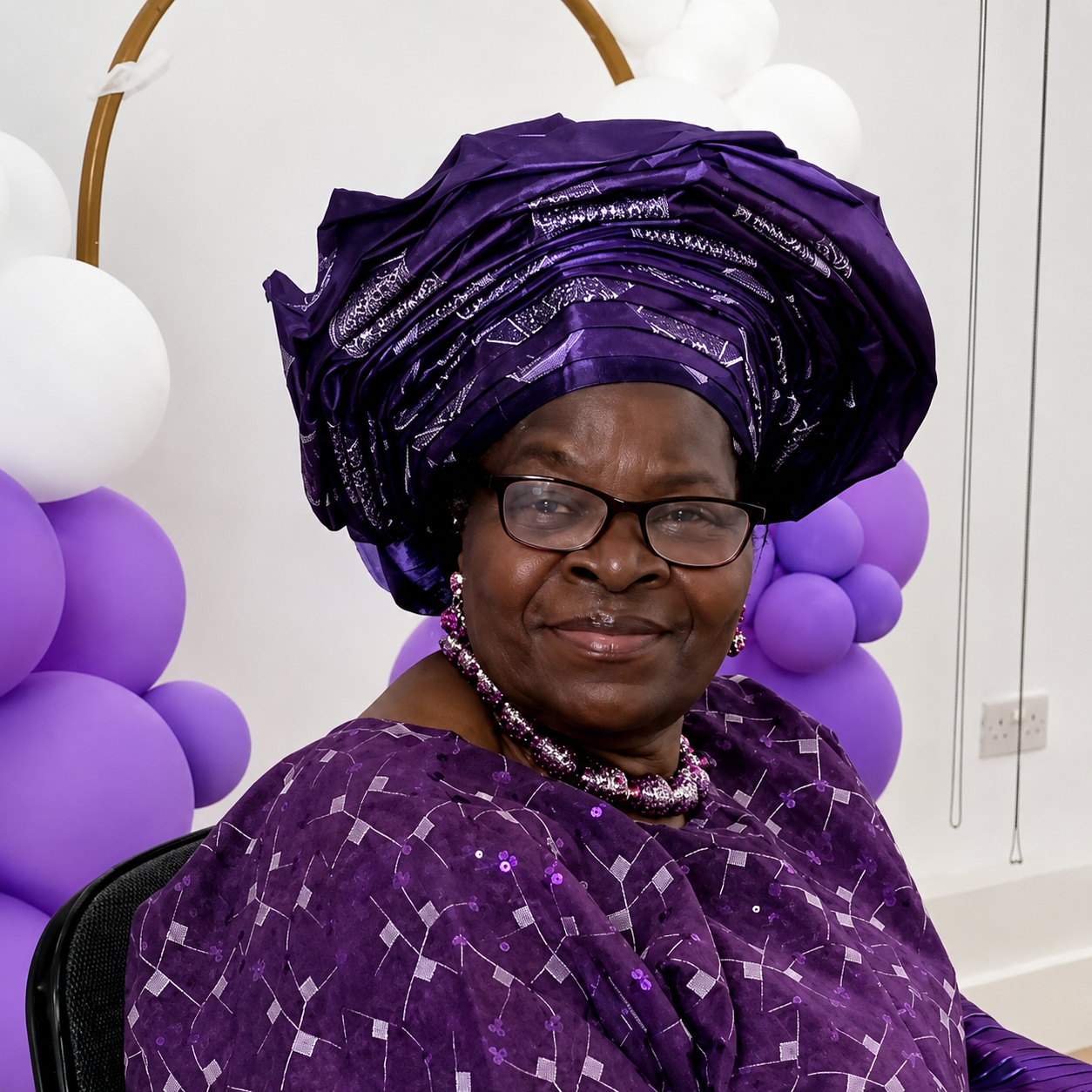 A woman with glasses, wearing traditional purple African attire and a headwrap, sitting in front of purple and white balloon decorations.