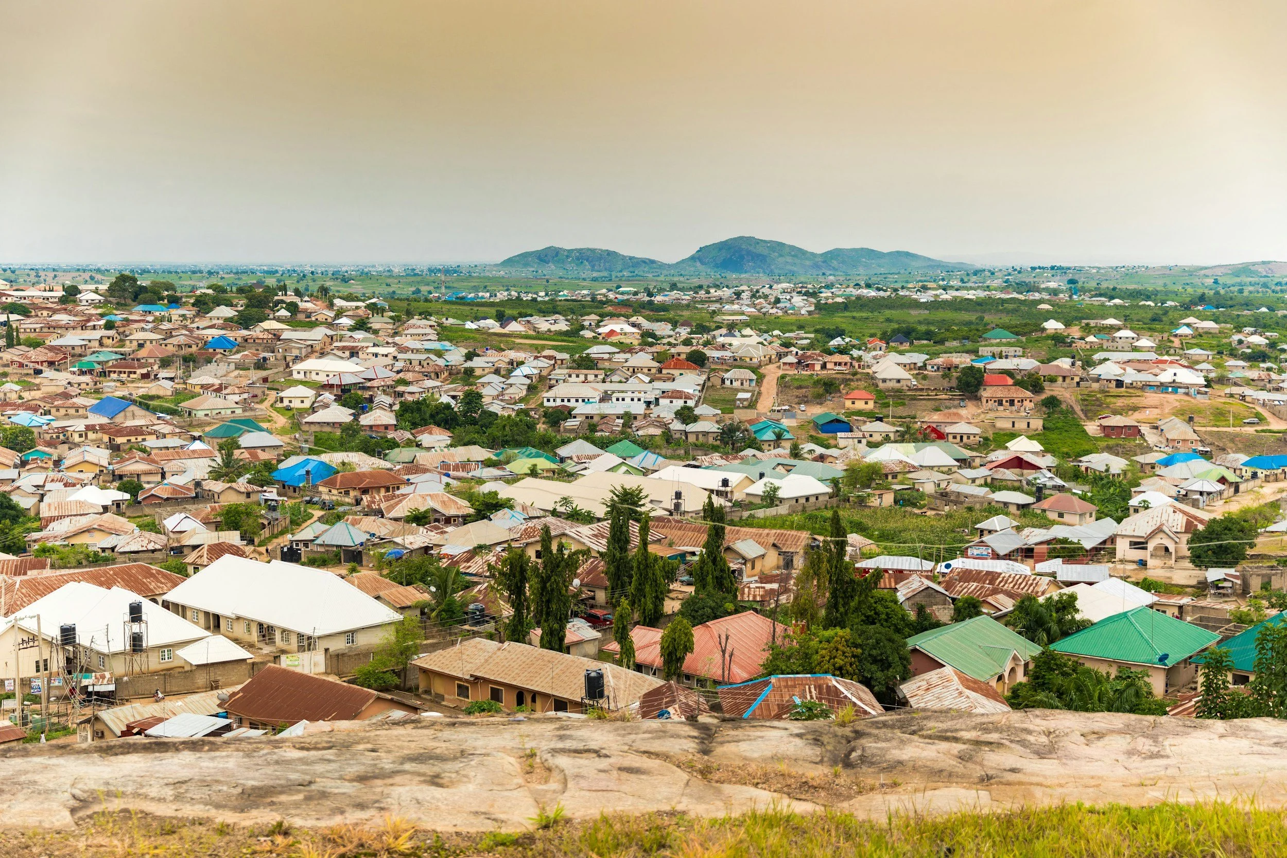 A sprawling view of a residential area with numerous houses featuring metal roofs, with some painted in bright colors, surrounded by greenery and trees. In the background, there are rolling hills under a hazy sky.