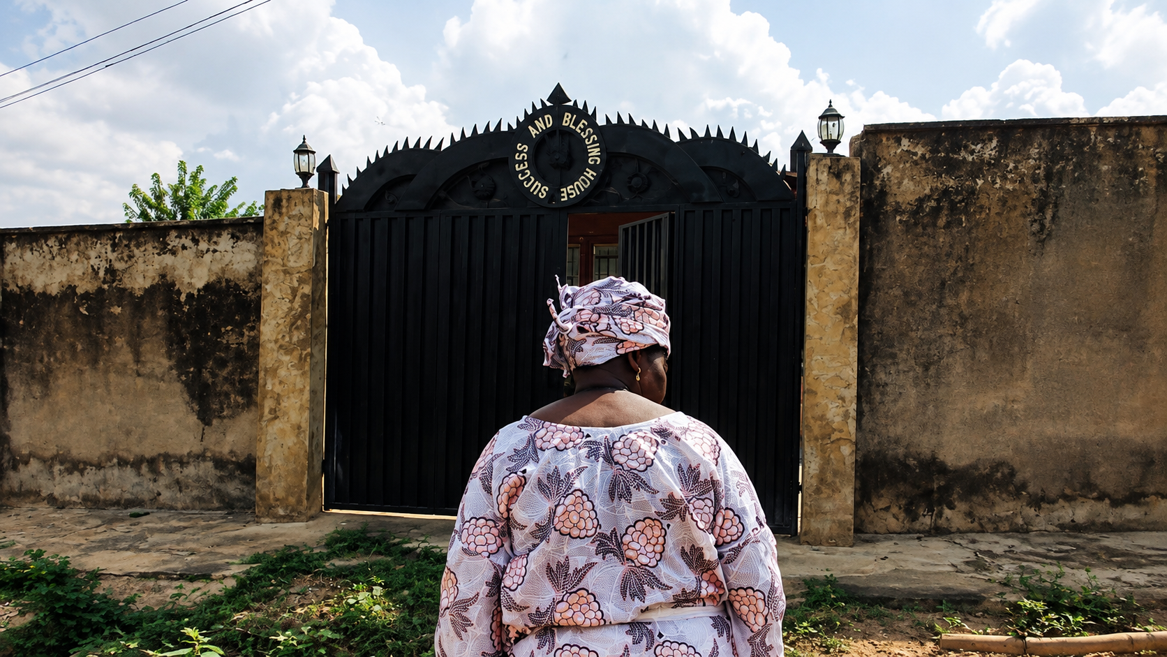 A woman in traditional patterned clothing and head wrap standing in front of a closed black gate with an inscription, flanked by weathered stone walls, under a partly cloudy sky.