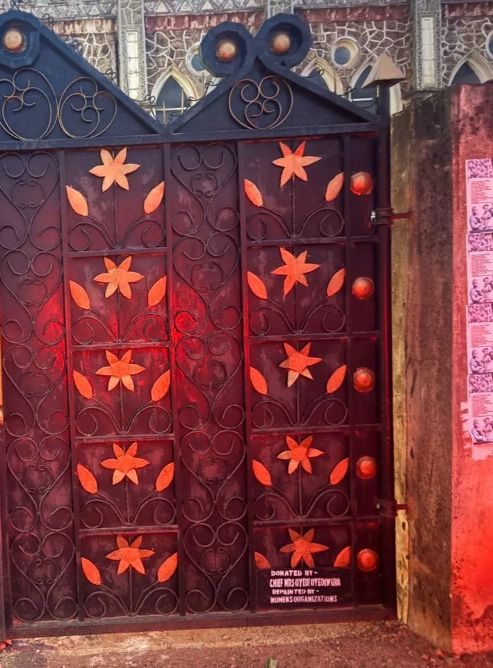 Decorative iron gate with orange flower and leaf patterns, at night, illuminated by red light, with signage indicating it was donated by a women's organization.
