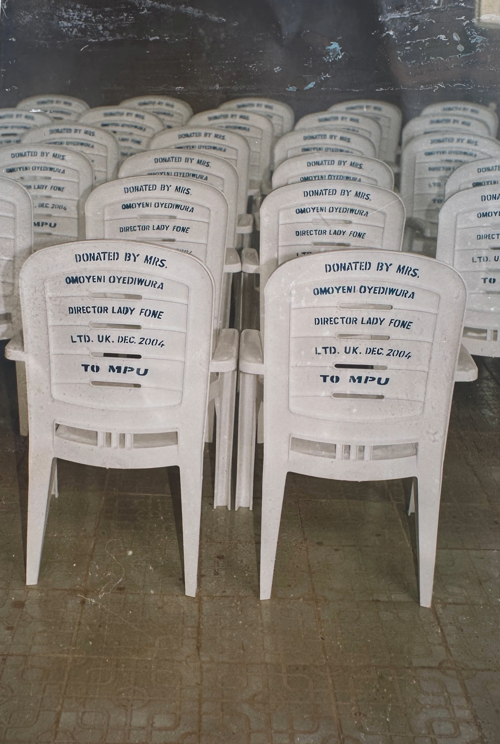 A group of white plastic chairs with text on the backrests that reads, 'Donated by Mrs. Omoyeni Oyediwura, Director Lady Fone, Ltd. UK Dec. 2006, to MPu.' The chairs are arranged in a semi-organized manner on a tiled floor with some dirt and marks, and they are seen through a glass window or door with reflections.