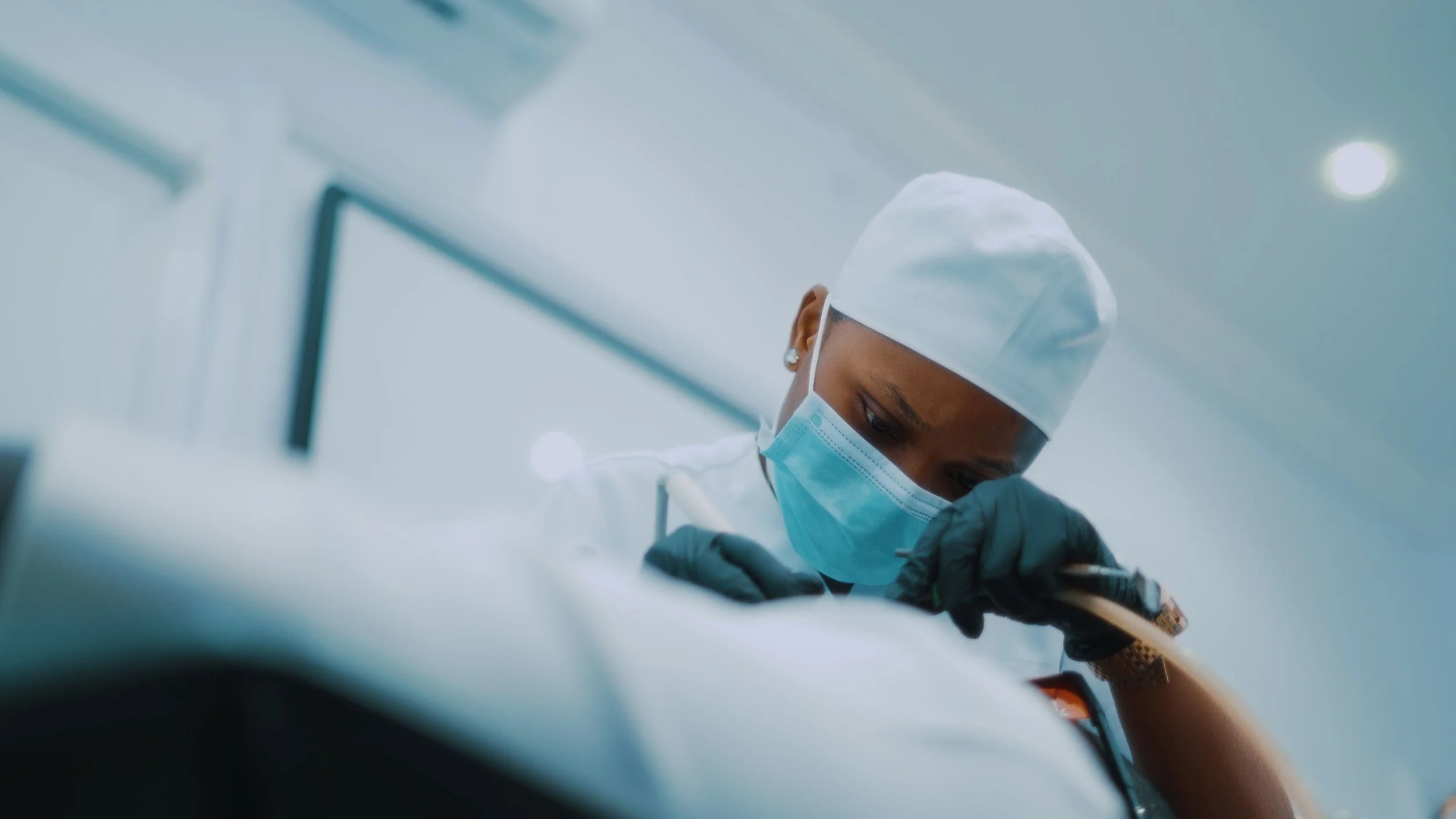 A healthcare worker wearing a surgical cap, face mask, and gloves, performing a medical procedure in a sterile environment.