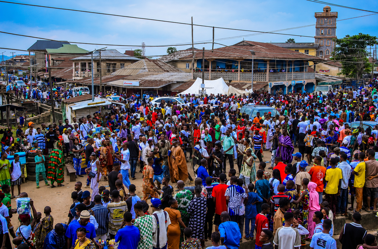 A crowded street scene in an urban area with many people, buildings, and vehicles, including cars and motorcycles.