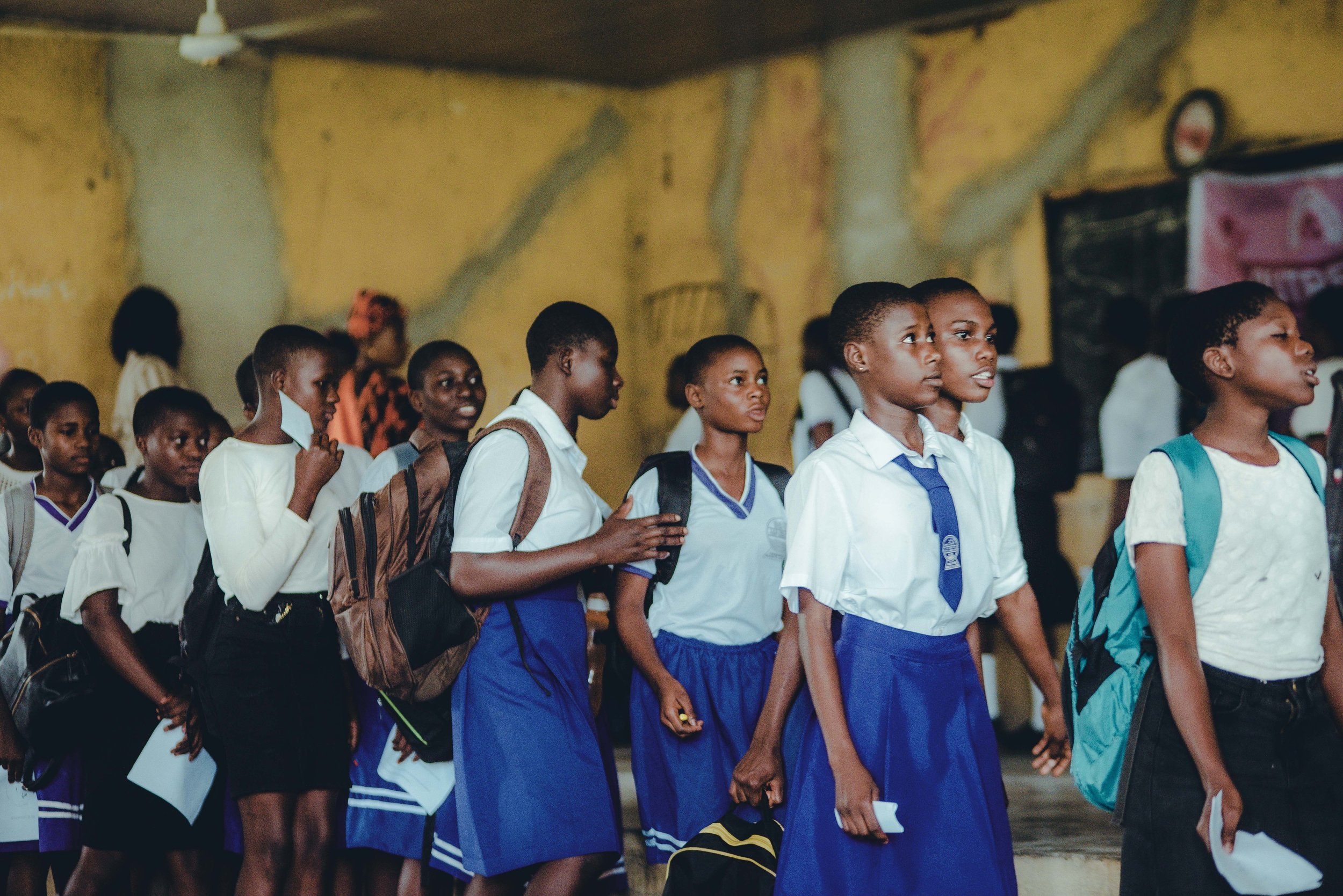 Group of school children wearing blue and white uniforms, standing in a classroom or hall, holding books and backpacks, some with facial masks hanging from their necks.