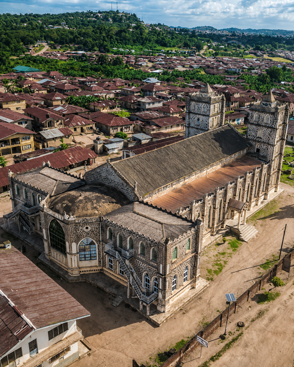 Aerial view of a historic church with two tall bell towers, a large main hall, and intricate stonework, surrounded by residential houses and greenery in a hilly landscape.