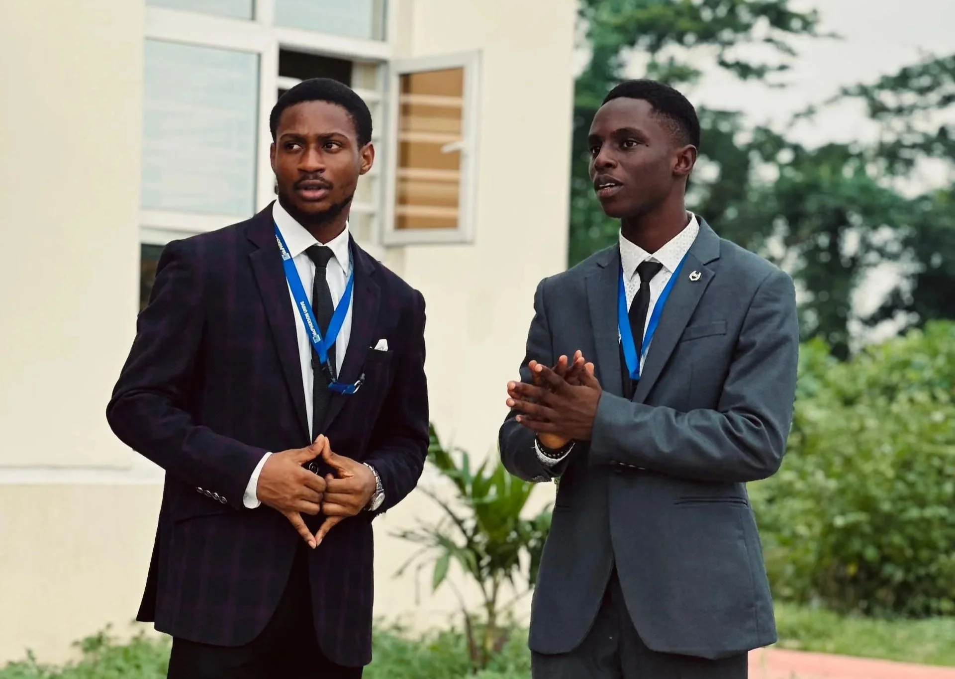 Two young men in suits and ties engage in a conversation outdoors in front of a building with open windows. One man gestures with his hands, while the other appears to be listening attentively. The scene is set outdoors with greenery visible in the background.