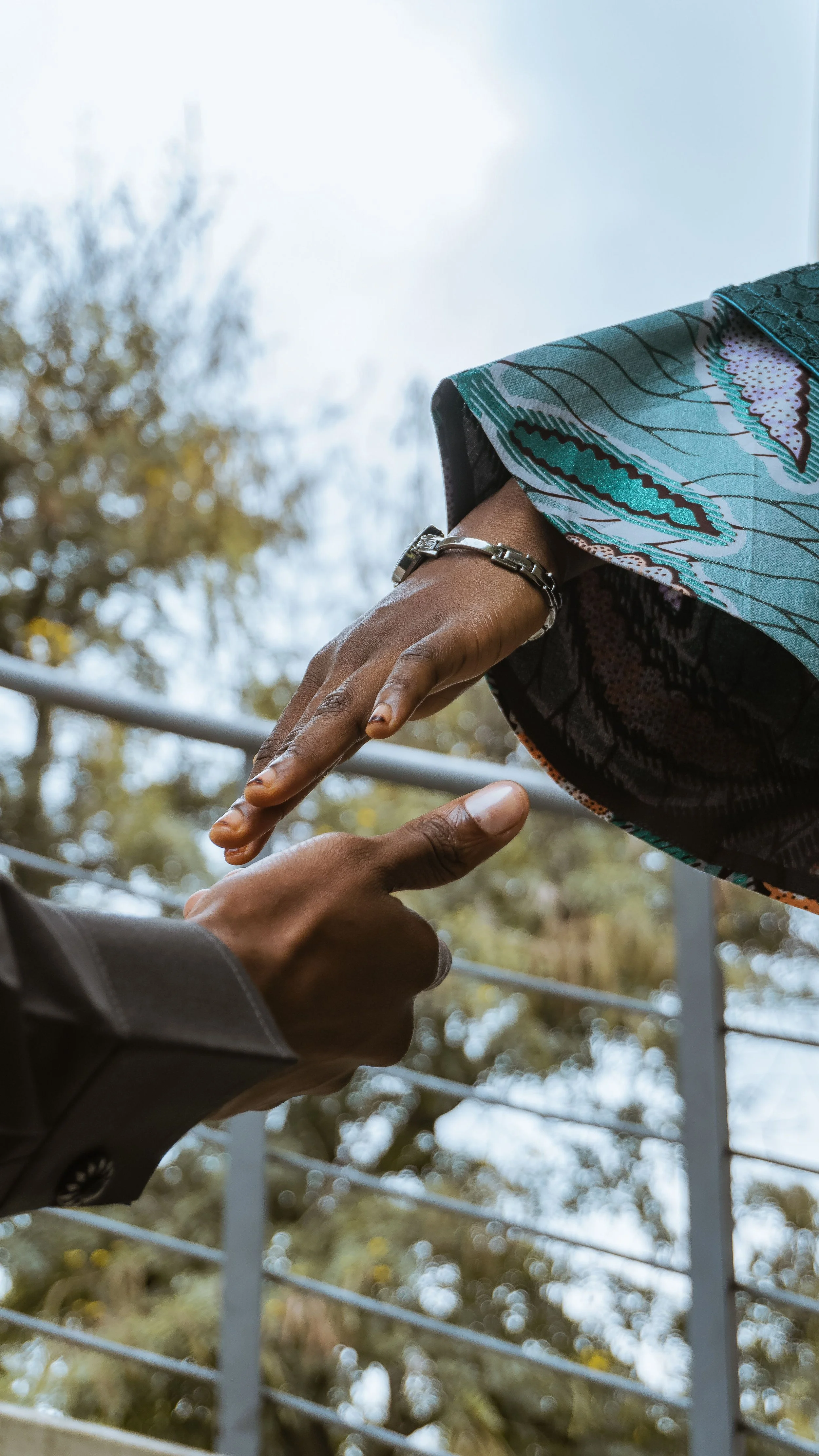Two people holding hands, with one person wearing a silver bracelet and a colorful patterned sleeve, outdoors near a metal fence and trees.