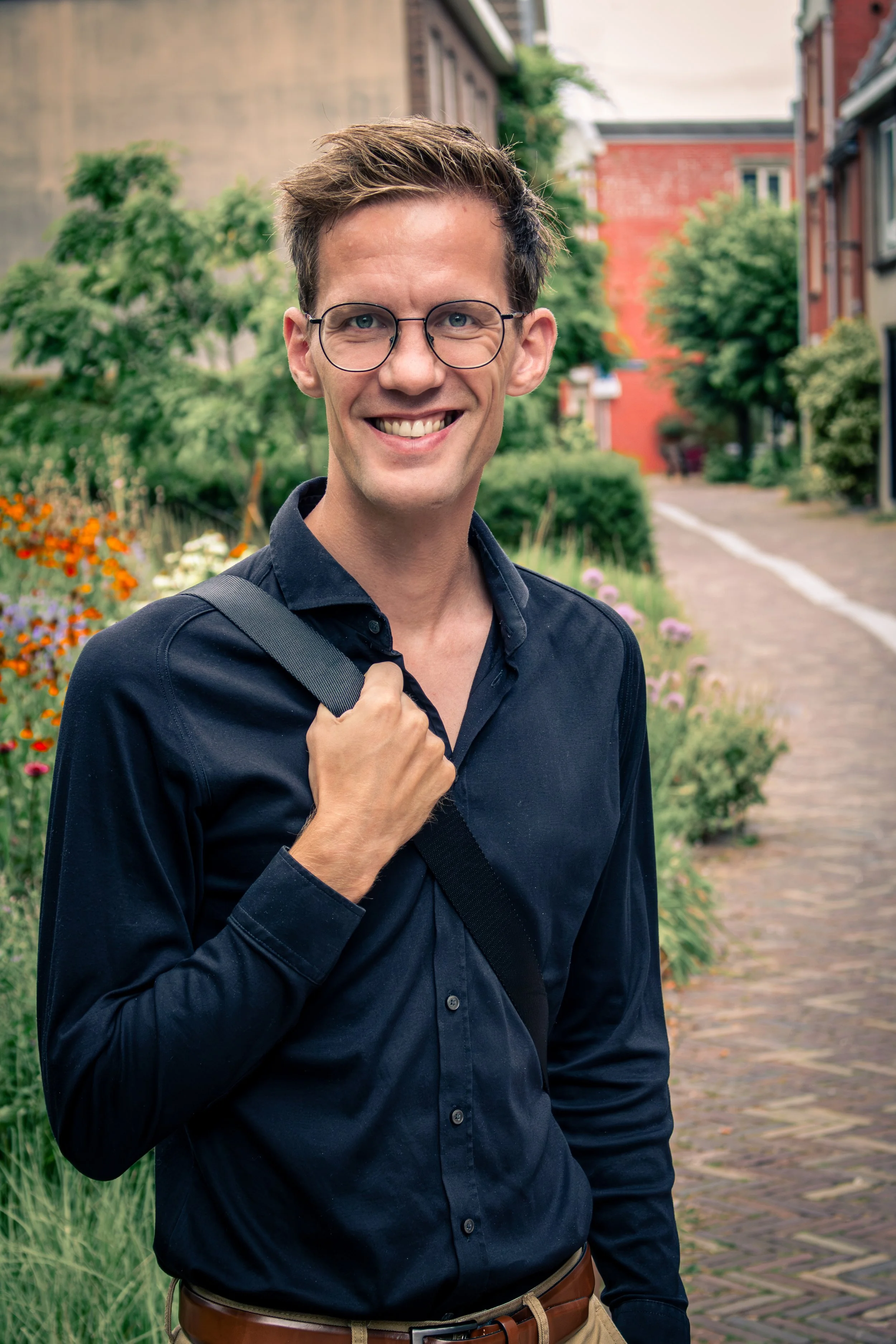 Man met bril en zwarte blouse lopend langs een smal zandpad met bloemen, groen struikgewas en oude gebouwen op de achtergrond.