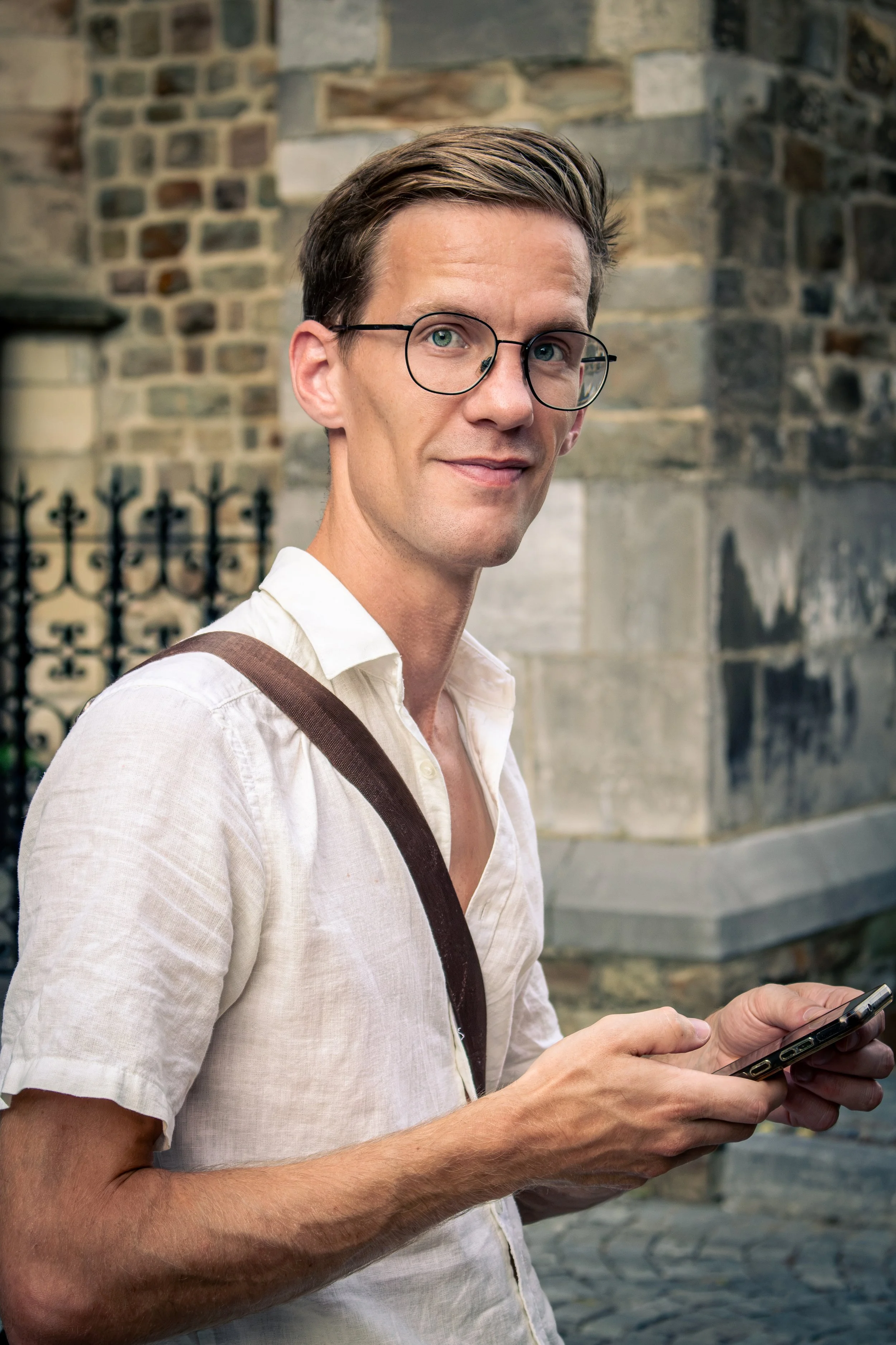 man met bril, witte blouse, en schoudertas die op straat met smartphone opereert, achtergrond van een stenen muur.