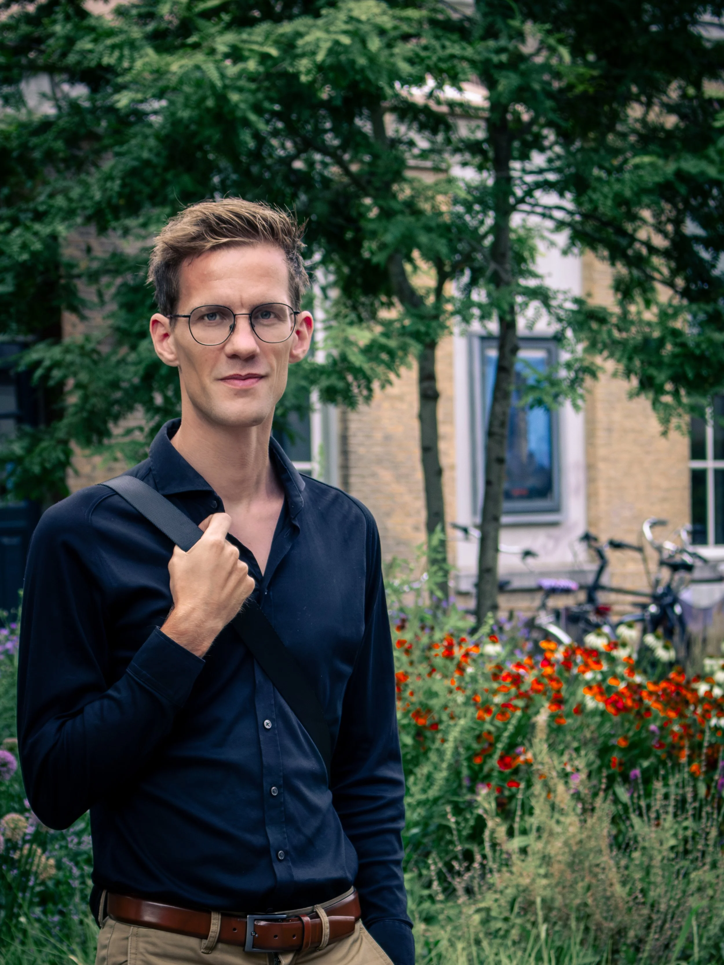 Een jonge man met bruin haar en een bril, gekleed in een donkerblauwe blouse en beige broeken, staat buiten voor een groene tuin met bloemen en bomen.
