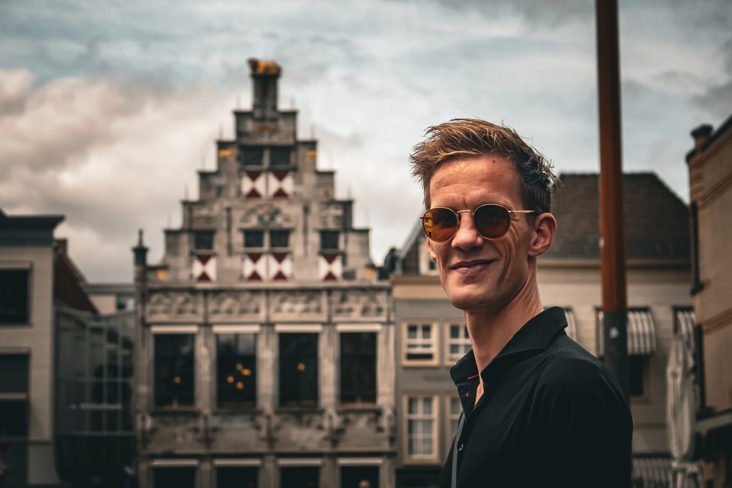 Close-up van een jongen met korte, lichtbruin haar, zonnebril en een zwarte jas, lachend op een straat met historische gebouwen.