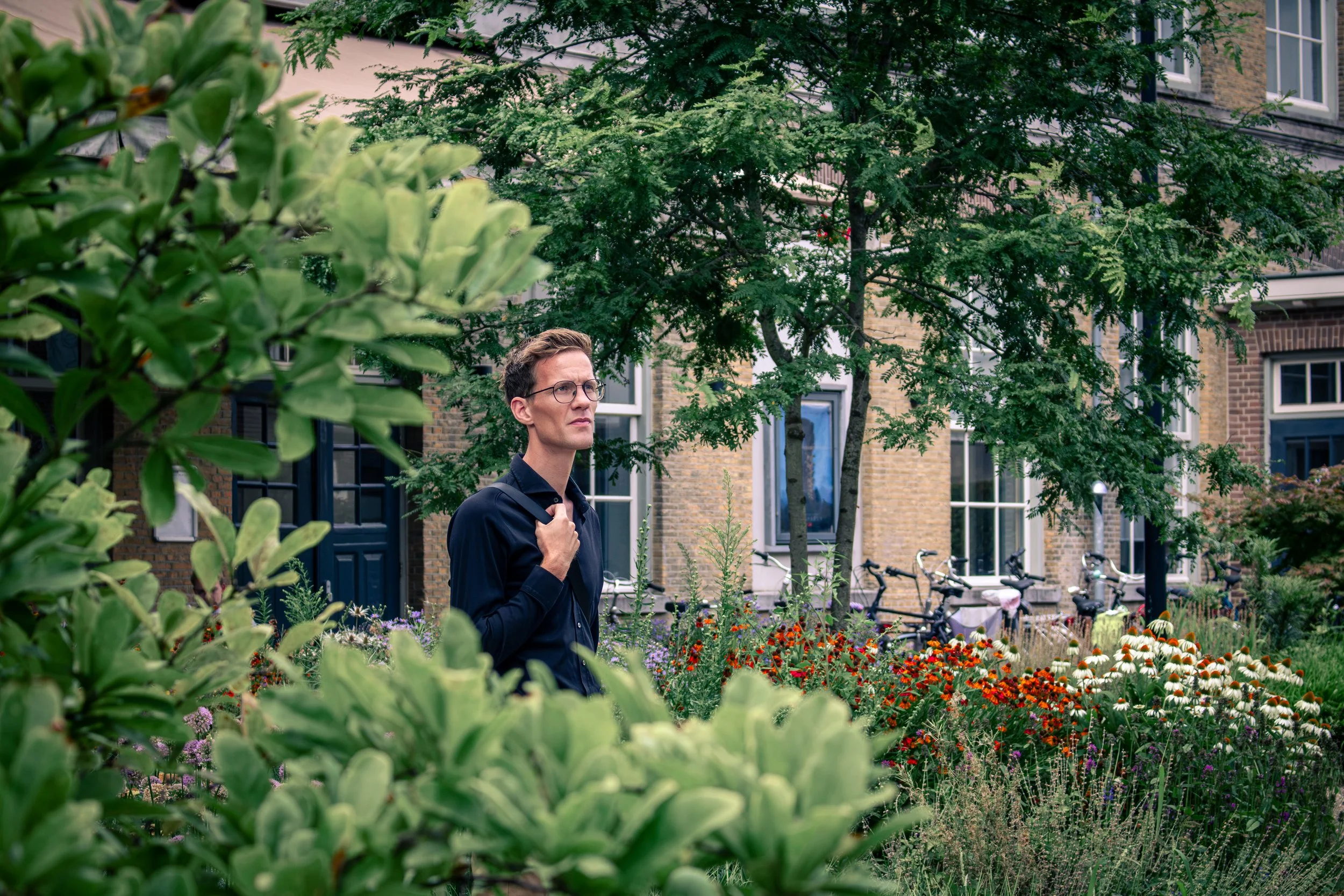 Man met bril en donker shirt loopt door een tuin met bloemen, planten en bomen in een stadswijk met historisch ogende gebouwen en fietsen op de achtergrond.