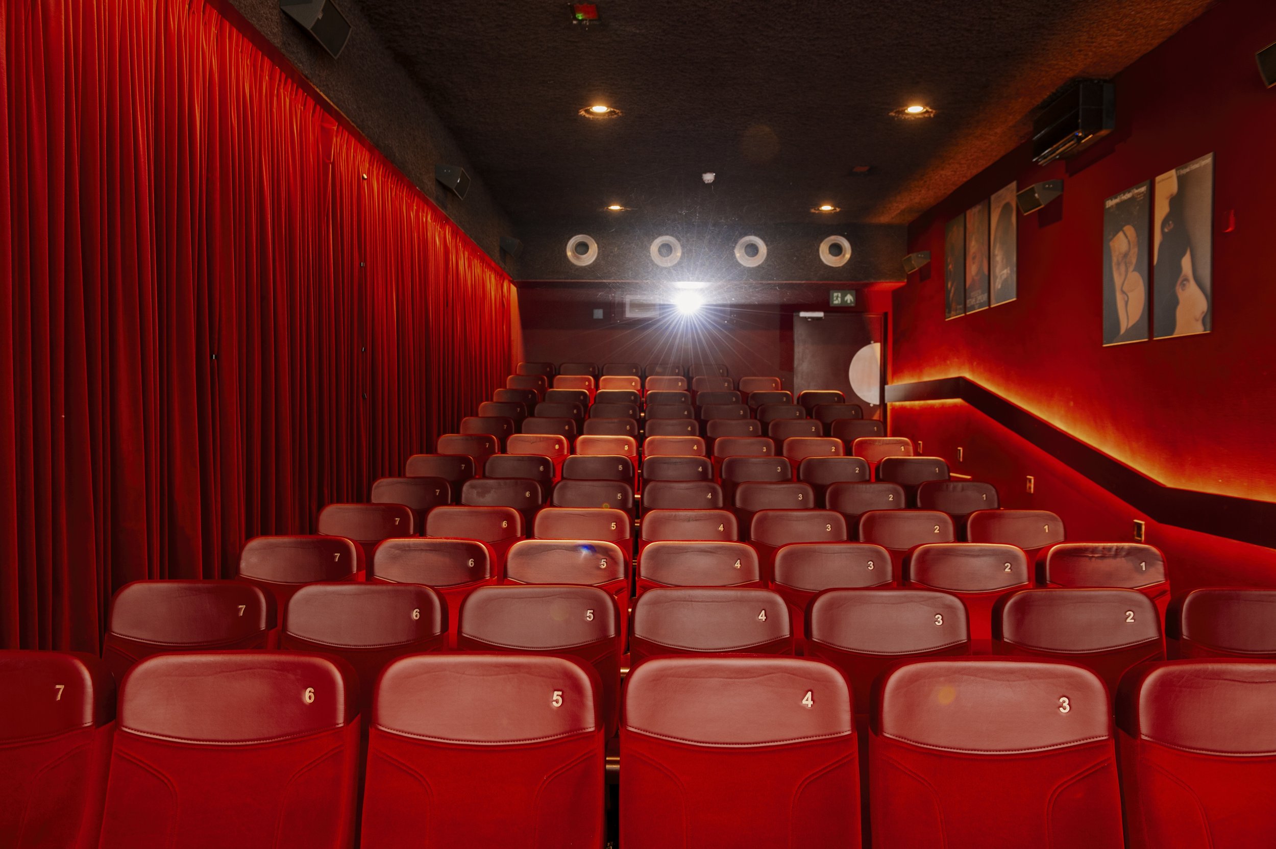 Inside a cinema with rows of red seats, a red curtain on the left, and movie posters on the right. Bright flash from the projector is visible at the back.