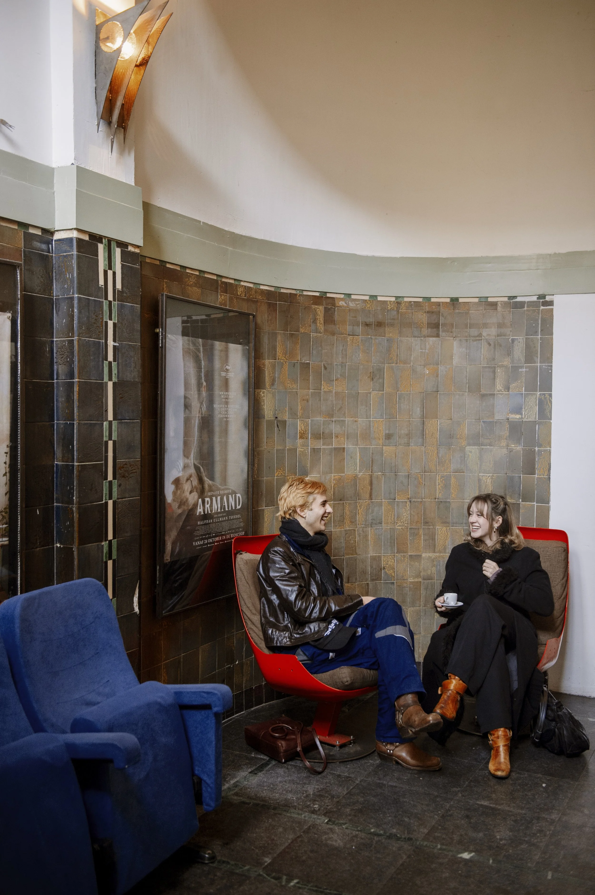 Two people sitting in red chairs talking and smiling, one holding a cup in a cozy cinema. There are blue cinema chairs in the foreground and tiled walls with artwork and a movie poster in the background.