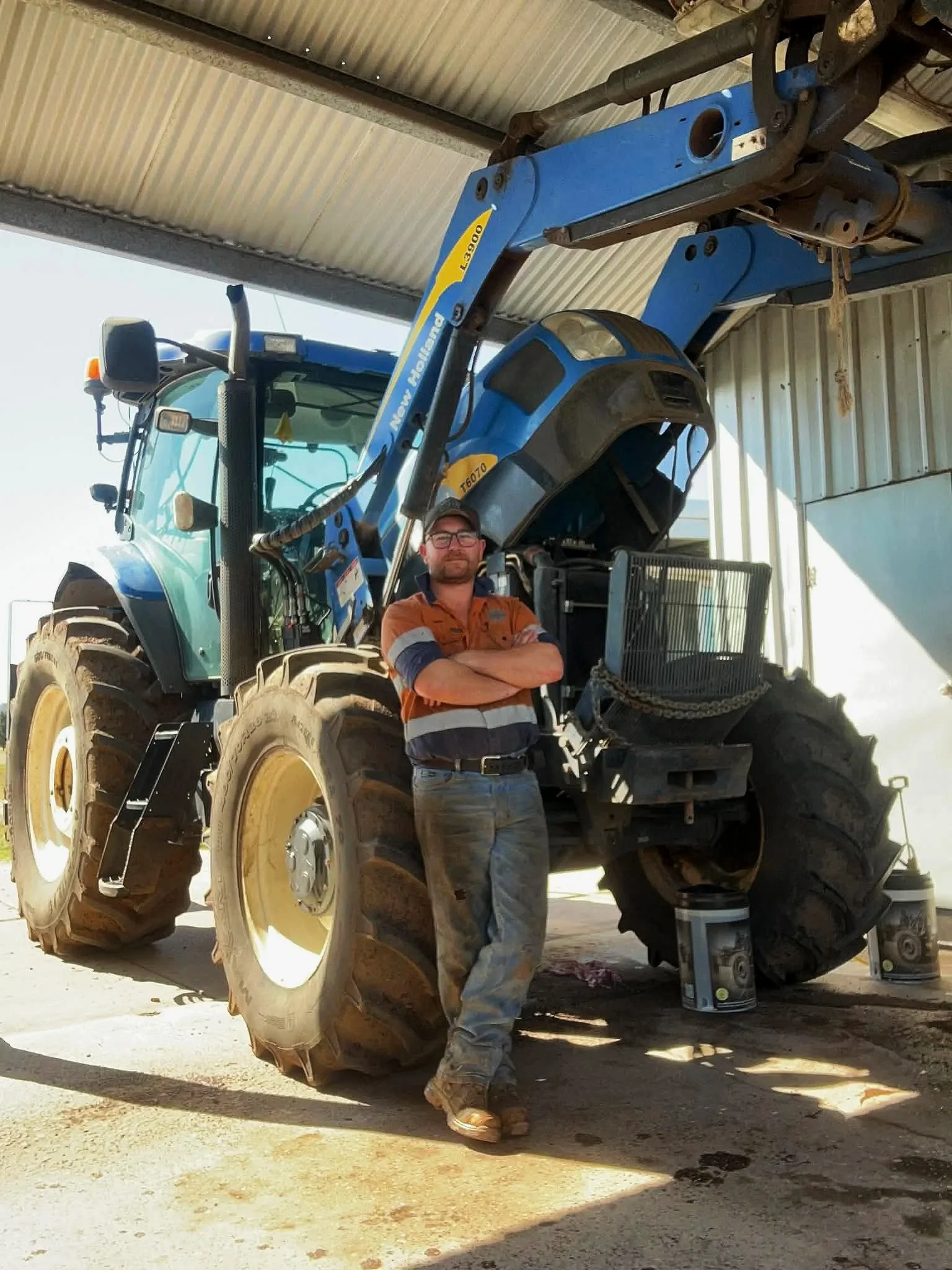 A man standing in front of a large blue tractor with yellow wheels, under a metal shed. The man, Chris, is a diesel mechanic specialising in field servicing and transport.