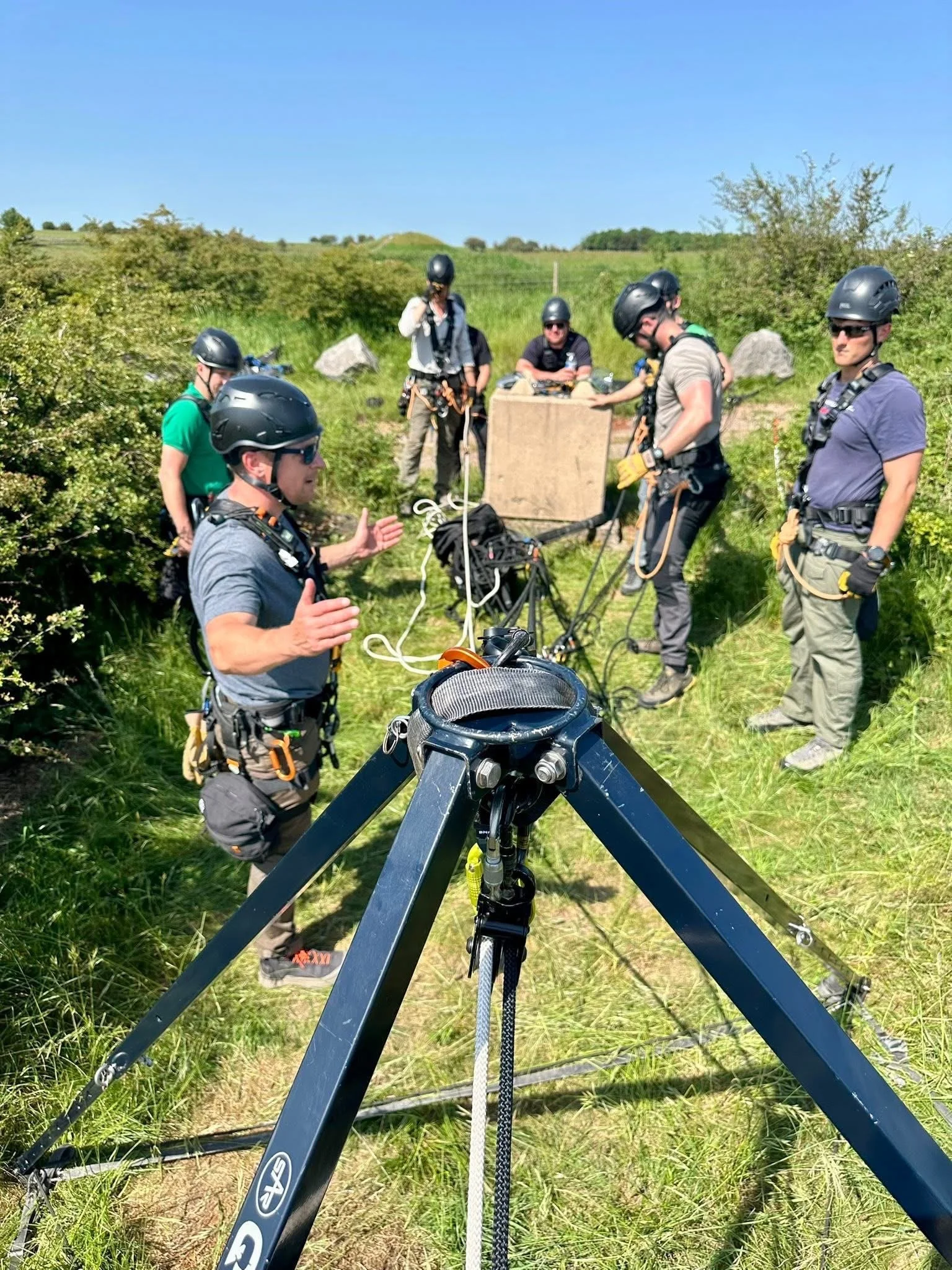 Rope rescue course delegates in helmets gathered around a tripod rescue system in a field