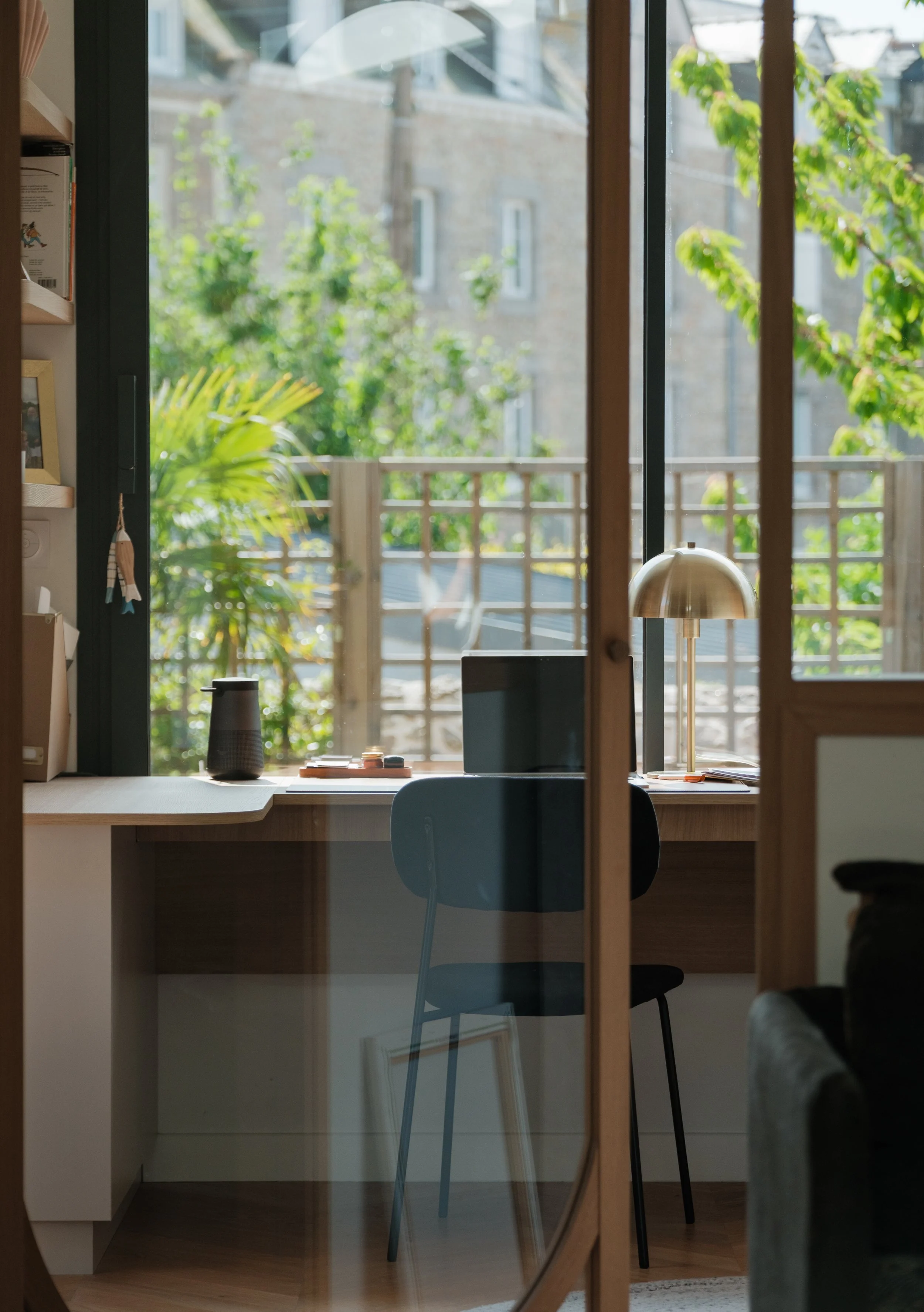 Espace de travail avec un bureau, un ordinateur portable, une lampe de bureau, une chaise bleue, et une fenêtre avec vue sur un balcon et des arbres en arrière-plan.