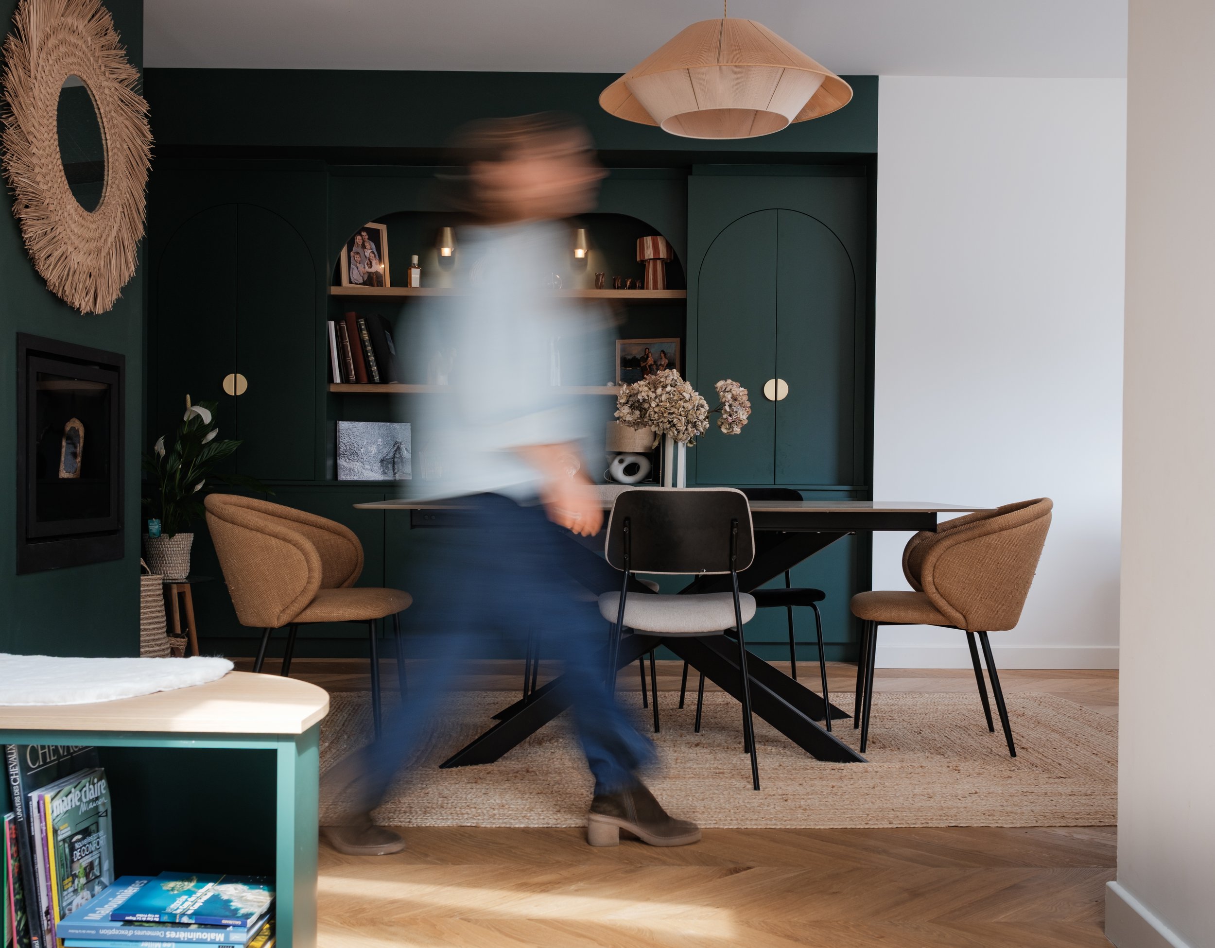 Un intérieur moderne avec une table en bois, des chaises en tissu beige, une bibliothèque verte foncée avec des objets décoratifs et des livres, un miroir en rotin au mur, et un lustre en bois suspendu au plafond. Une personne marche rapidement dans la pièce, apparaissant floue.