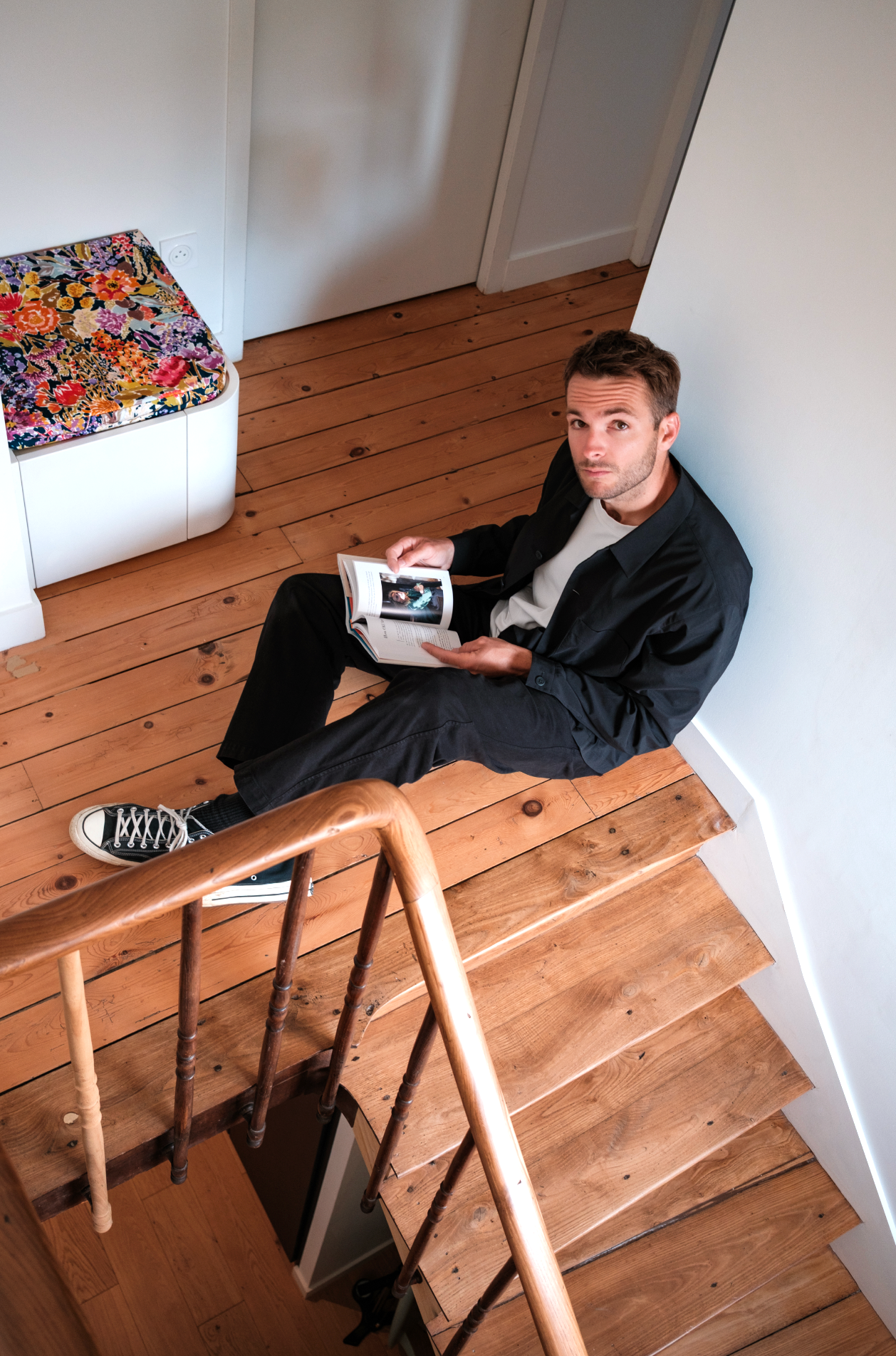 Jeune homme assis par terre dans une maison, lisant un livre, avec un escalier en bois devant lui, murs blancs, sol en bois, porte blanche et un meuble avec un coussin coloré.
