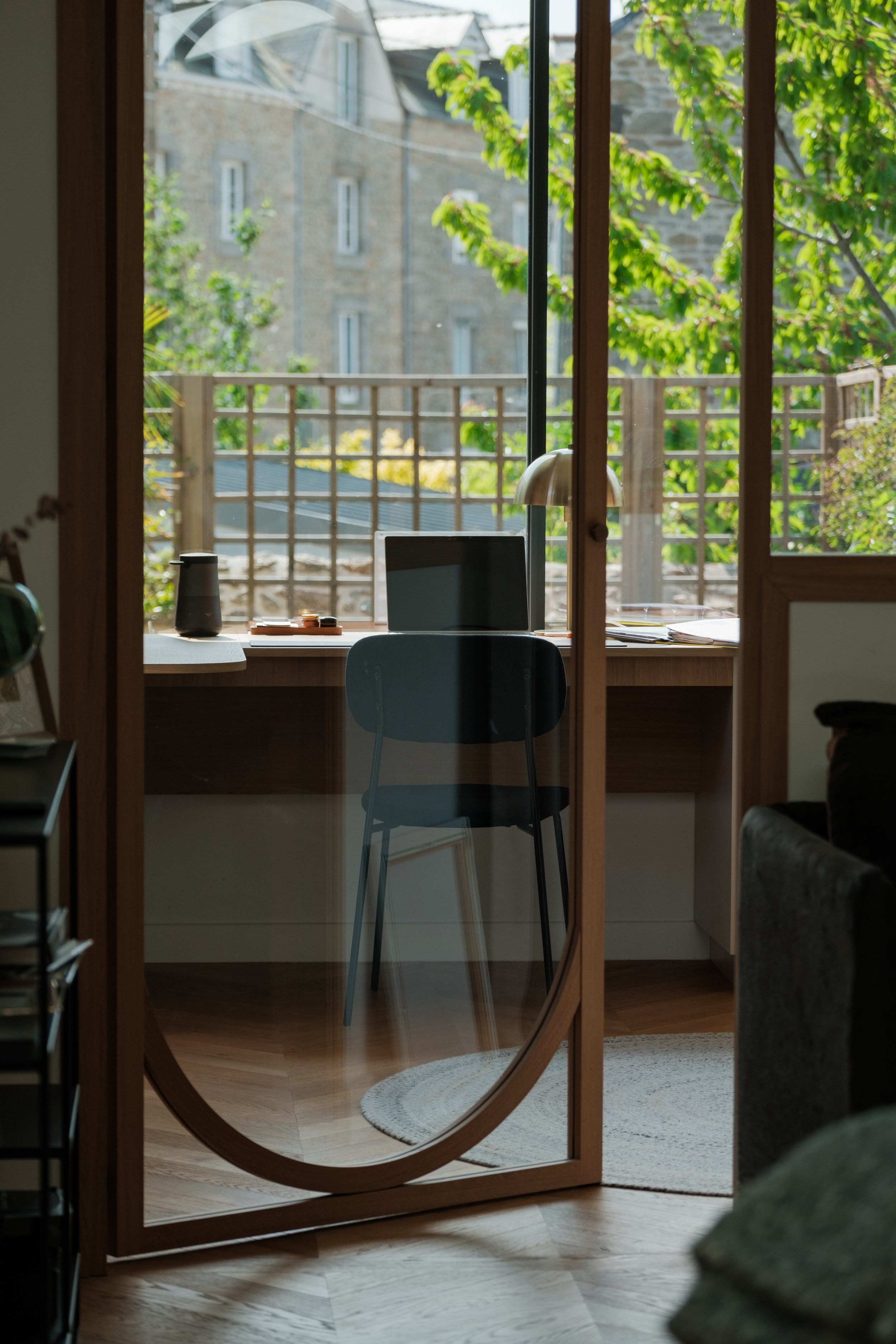 Vue à travers une porte en verre montrant un bureau avec un ordinateur portable, une lampe, une chaise, et un balcon avec des arbres et un bâtiment en pierre en arrière-plan.