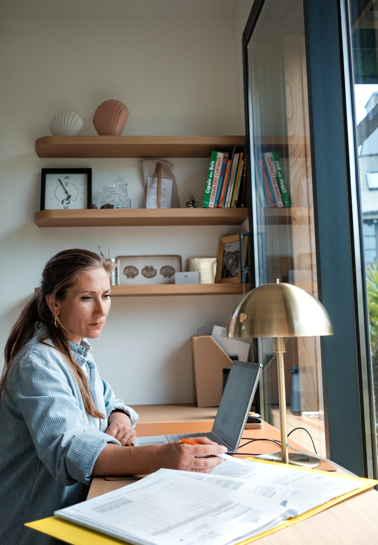 Une femme assise à un bureau utilise un ordinateur portable, avec des documents étalés à l'avant, dans une pièce bien éclairée par une grande fenêtre. Sur la table se trouve une lampe en laiton.