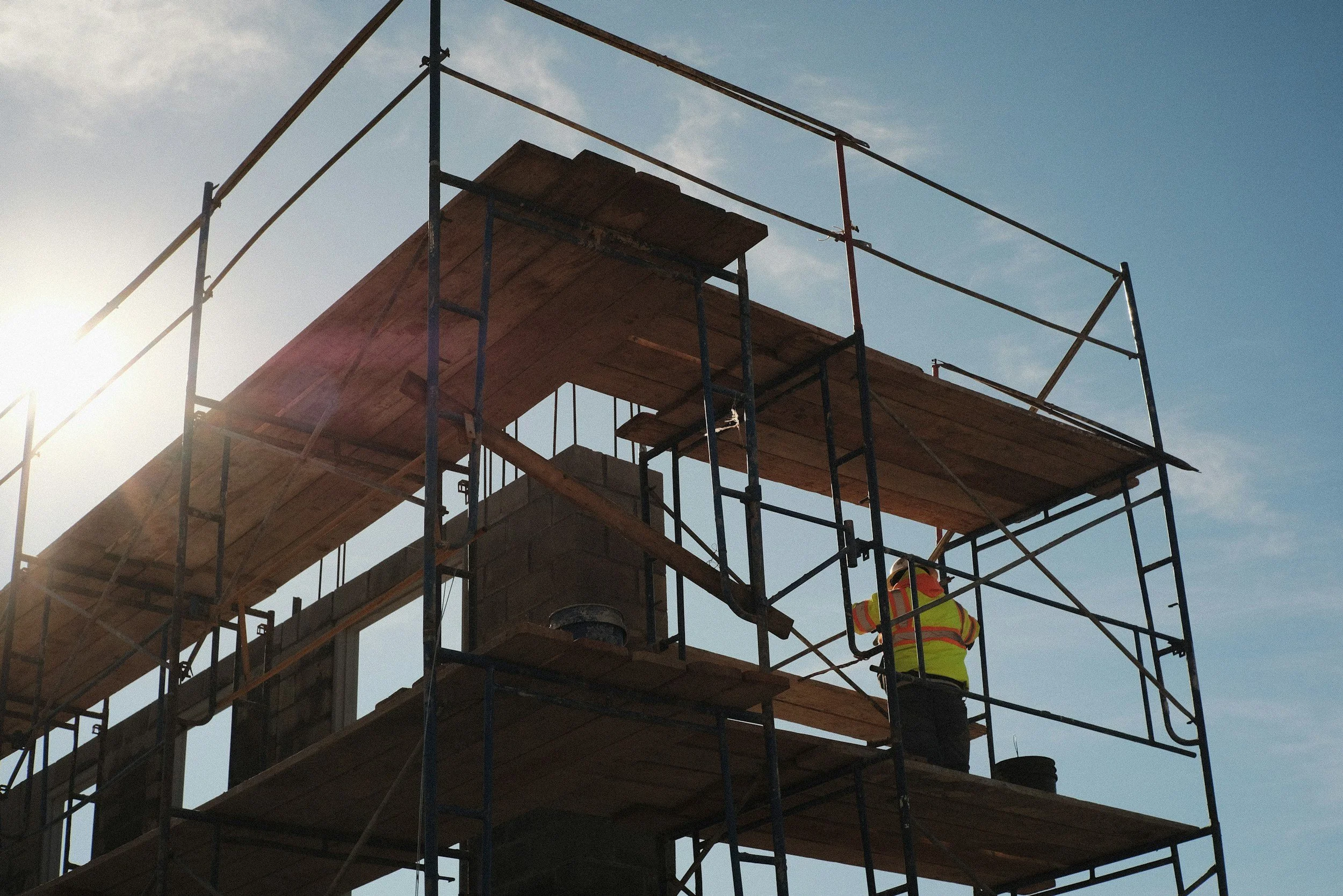Construction worker in safety vest and helmet working on a wooden scaffolding structure at a building site against a blue sky.