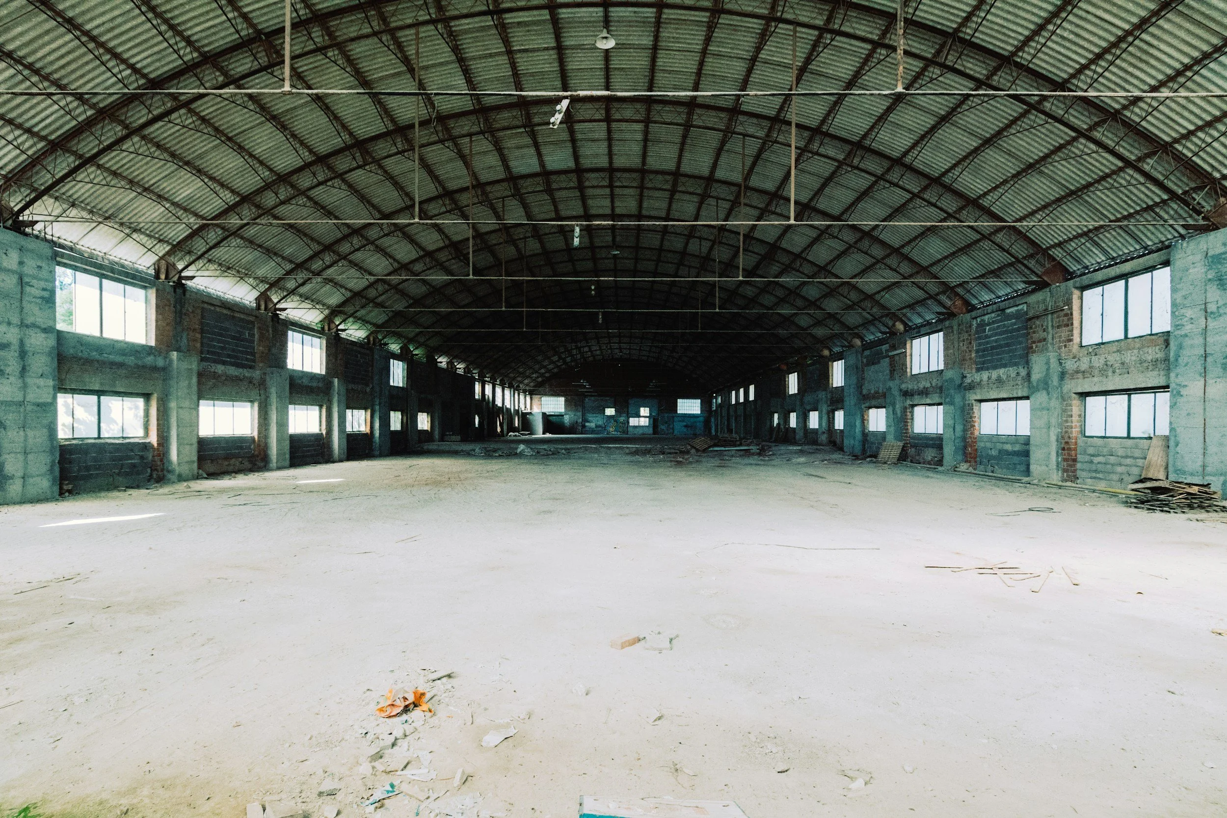 An empty, unfinished indoor space, possibly a warehouse or gym, with a high arched metal roof, large windows on the sides, and a concrete floor with some debris.